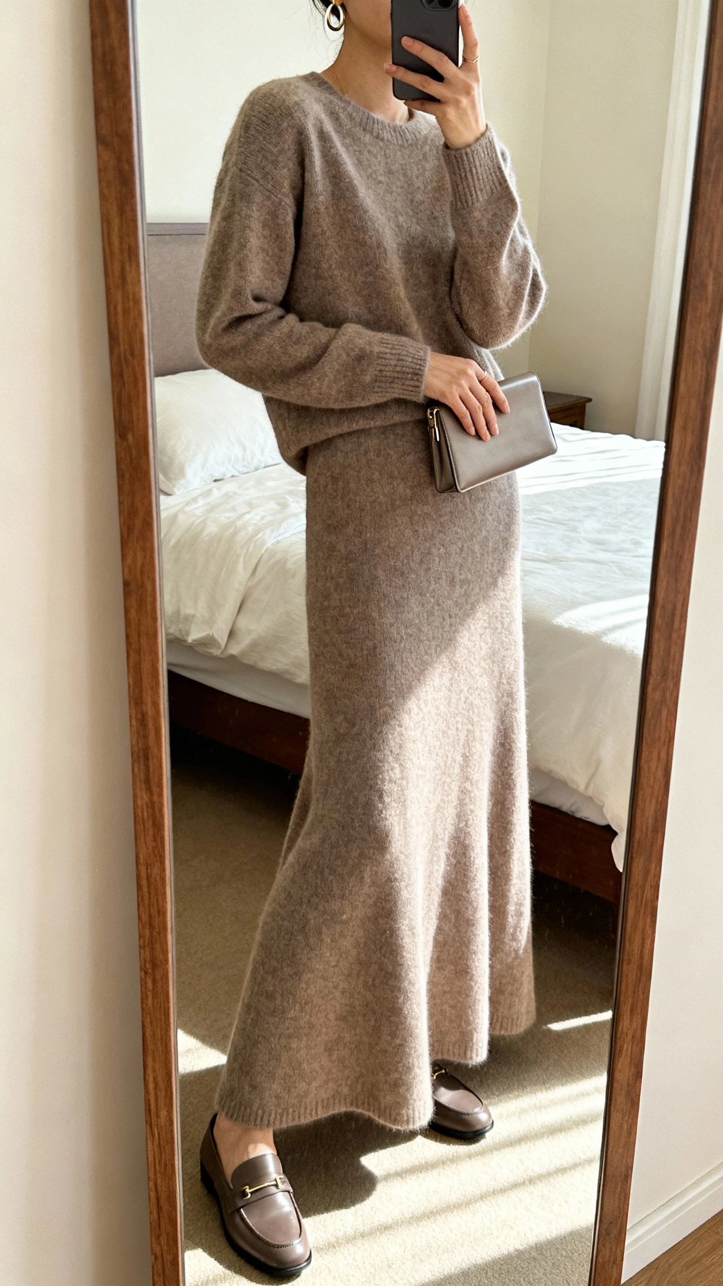iPhone mirror selfie of a woman wearing a tonal knit set: long-sleeve knit top and matching sweeping long skirt in the same shade, streamlined loafers, subtle hoop earrings, and a slim clutch, face not visible, side angle, bedroom full-length mirror, soft morning light, iPhone photo quality.