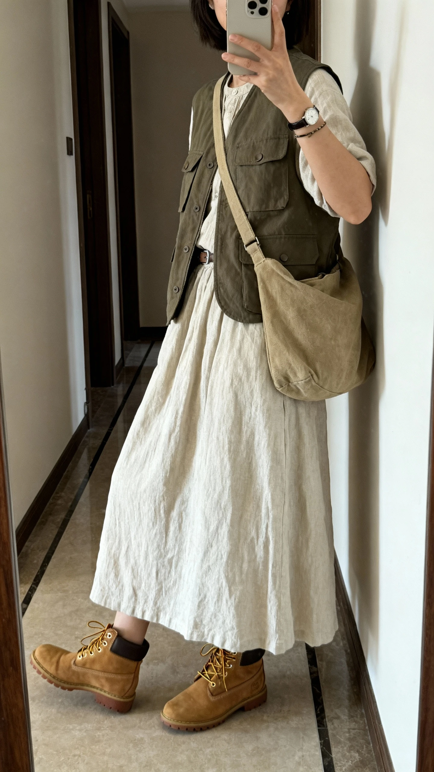 iPhone mirror selfie of a woman wearing a utility vest over a breathable cotton maxi dress with lace-up boots, canvas crossbody and a simple bracelet watch, face not visible, side angle, entryway full-length mirror, natural morning light, iPhone photo quality