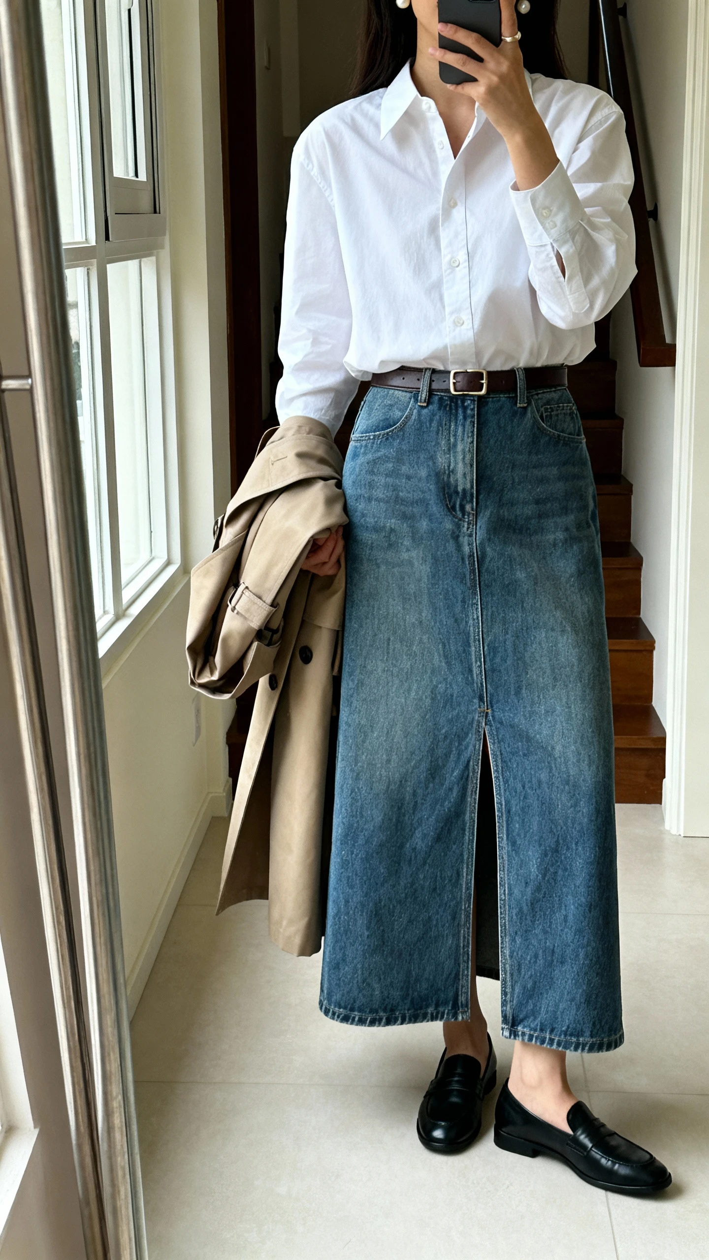 iPhone mirror selfie of a woman wearing a vintage-inspired denim maxi skirt with front slit, tucked crisp white button-down, black loafers, slim leather belt, pearl studs, and a structured trench draped over arm, face not visible, cropped at neck, entryway full-length mirror with natural window light, iPhone photo quality.