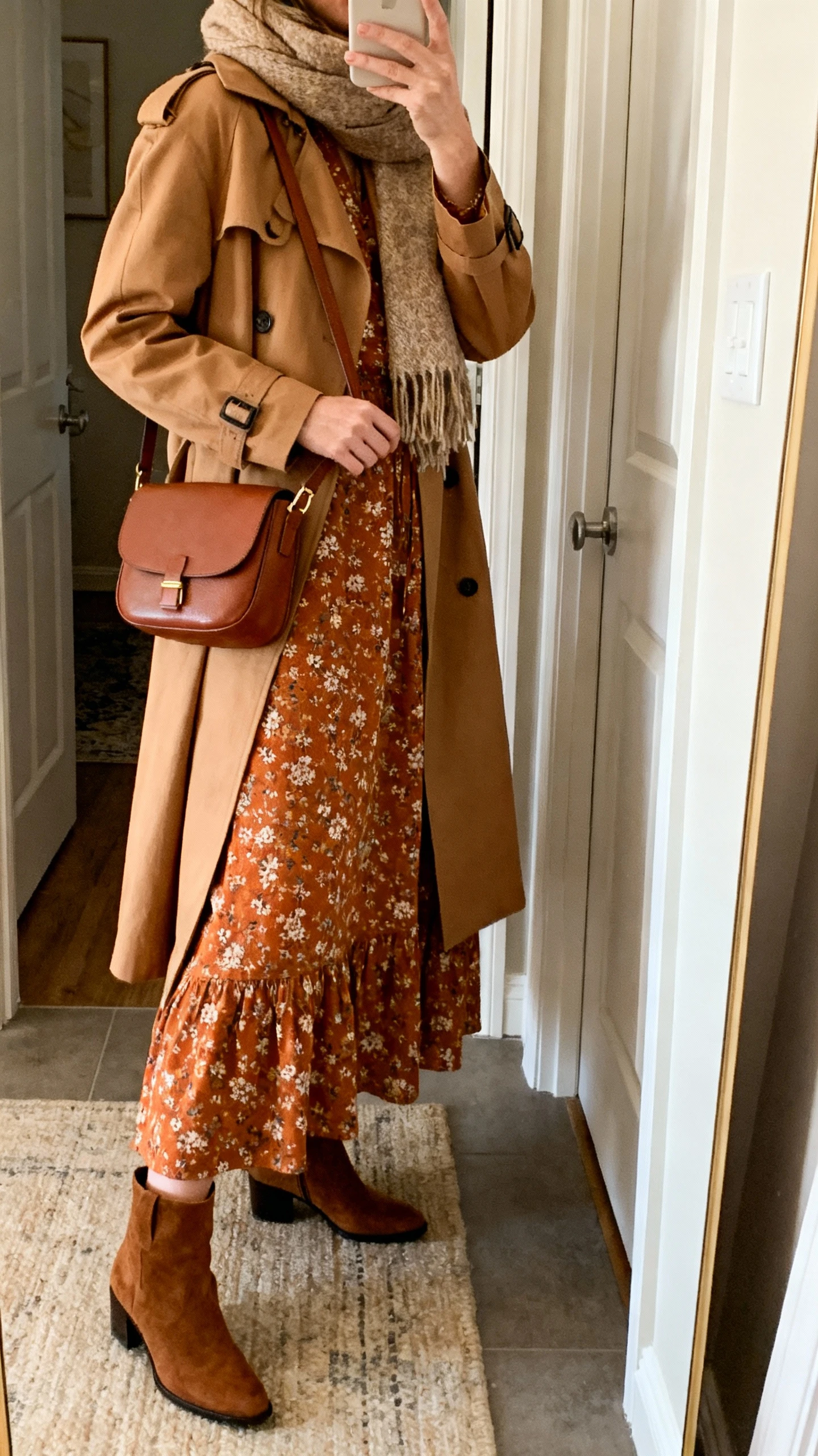 iPhone mirror selfie of a woman wearing a warm-toned autumn floral cotton maxi dress with a camel trench coat, ankle boots, a brown crossbody bag, and a cozy scarf, face not visible, side angle, entryway mirror, soft indoor lighting, iPhone photo quality.