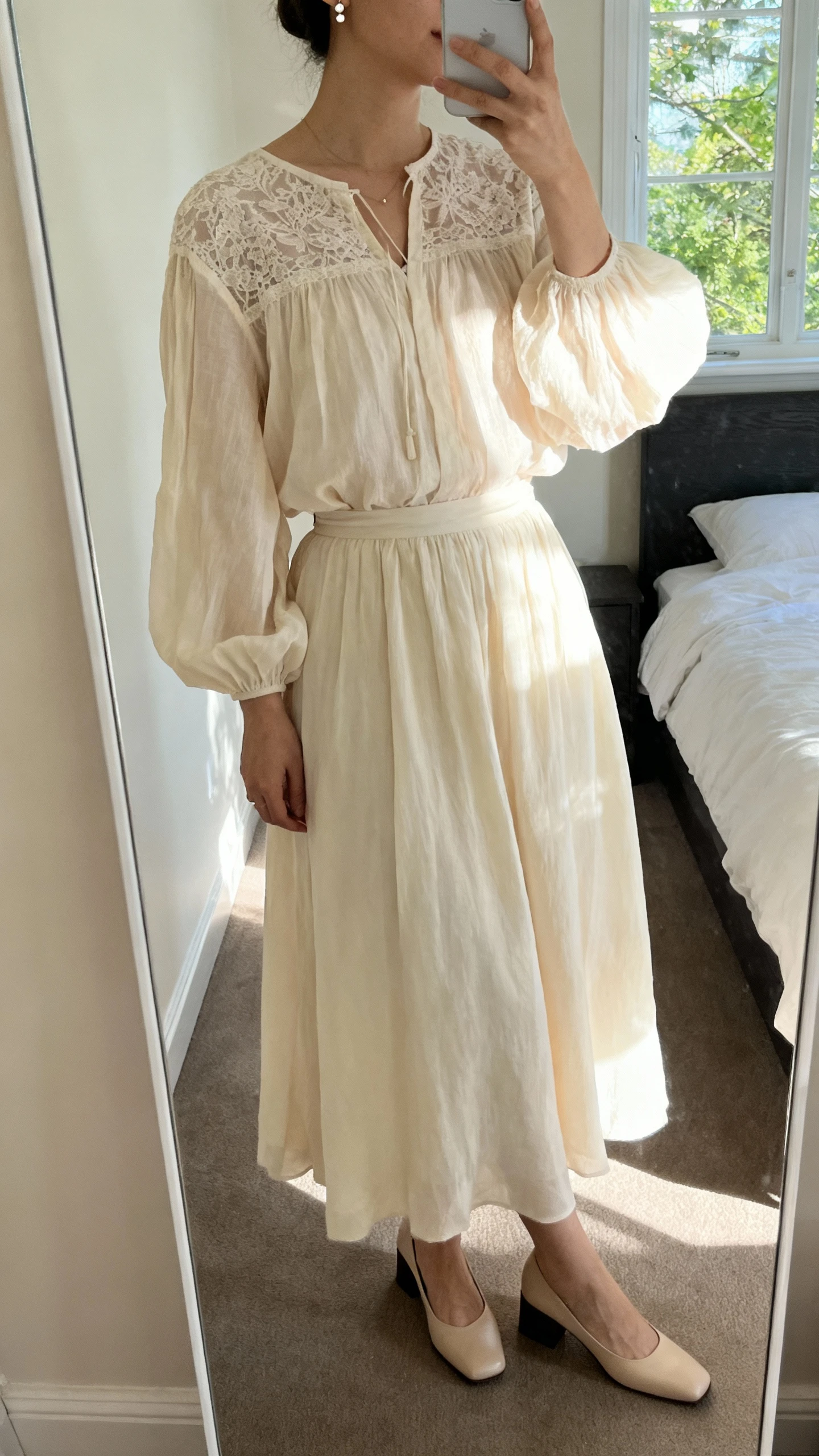 iPhone mirror selfie of a woman wearing a whisper-lace poet blouse with billowy sleeves tucked into a flowing midi skirt, paired with delicate stud earrings and low block-heel pumps, face not visible, cropped at neck, bedroom mirror with natural morning window light, iPhone photo quality.