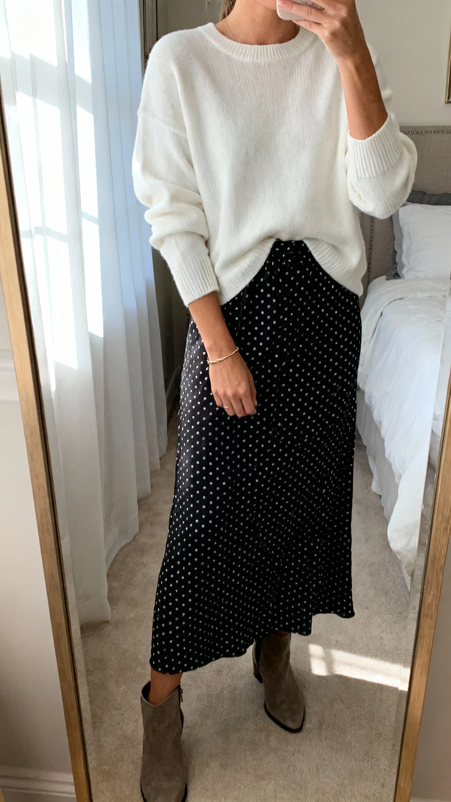 iPhone mirror selfie of a woman wearing a white crewneck knit sweater layered over a black polka dot maxi dress (skirt visible), waist slightly tucked, ankle boots and delicate bracelet, face not visible, cropped at neck, bedroom mirror, soft window light, iPhone photo quality.