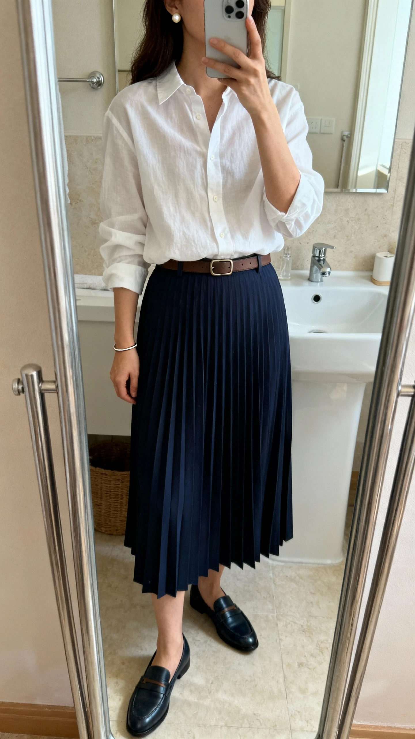 iPhone mirror selfie of a woman wearing a white slightly relaxed button-down shirt tucked into a navy pleated midi skirt, classic loafers, a minimalist leather belt, quiet pearl studs and a thin bracelet, face not visible, bathroom mirror, morning indoor lighting, iPhone photo quality.