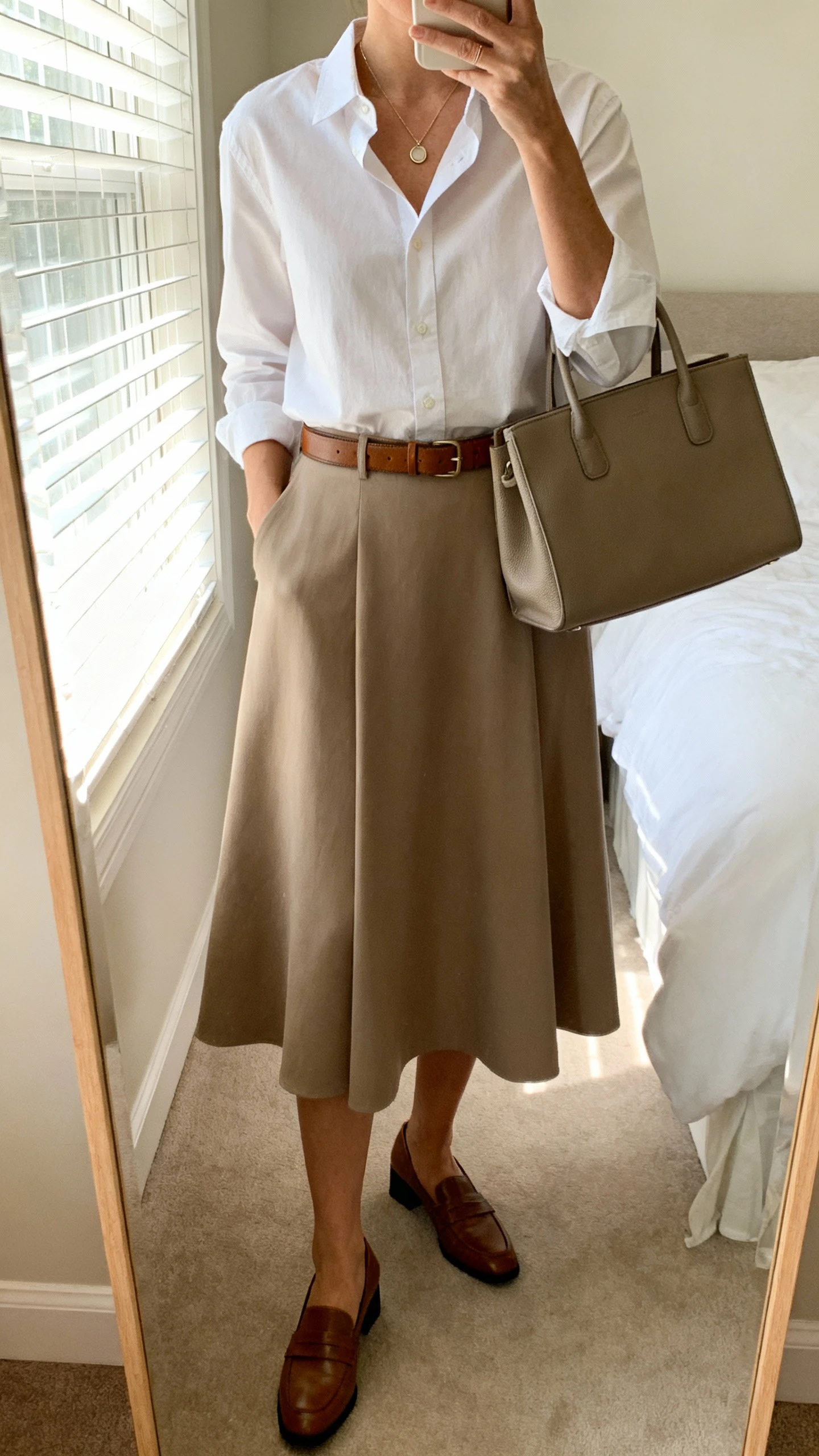 iPhone mirror selfie of a woman wearing an A-line midi skirt, crisp tucked-in button-up shirt, slim leather belt, low block-heel loafers, delicate pendant necklace, and a structured tote, face not visible, cropped at neck, bedroom mirror, natural window light, iPhone photo quality.