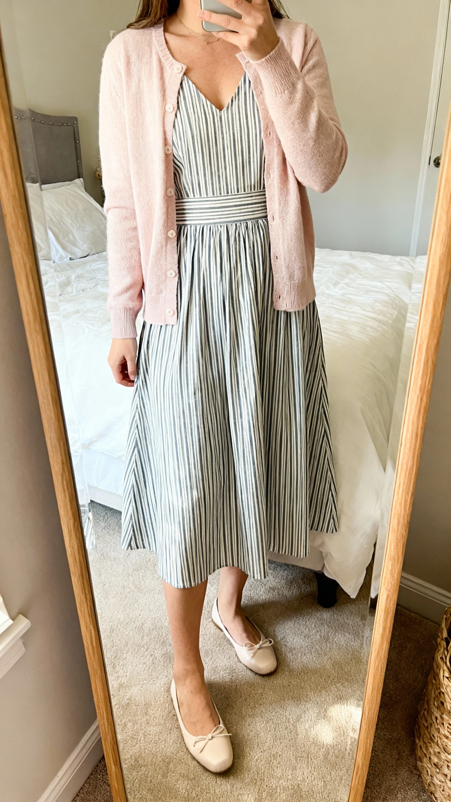 iPhone mirror selfie of a woman wearing an A-line striped dress layered with a pastel cardigan and classic ballet flats, face not visible, cropped at shoulders, cozy bedroom mirror with soft daylight, iPhone photo quality.