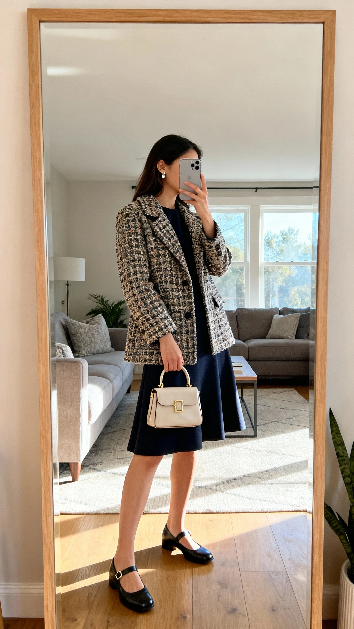 iPhone mirror selfie of a woman wearing an A-line dress with Mary Janes and a statement coat, carrying a small top-handle bag and wearing dainty earrings, face not visible, side angle, living room full-length mirror, soft morning window light, iPhone photo quality.