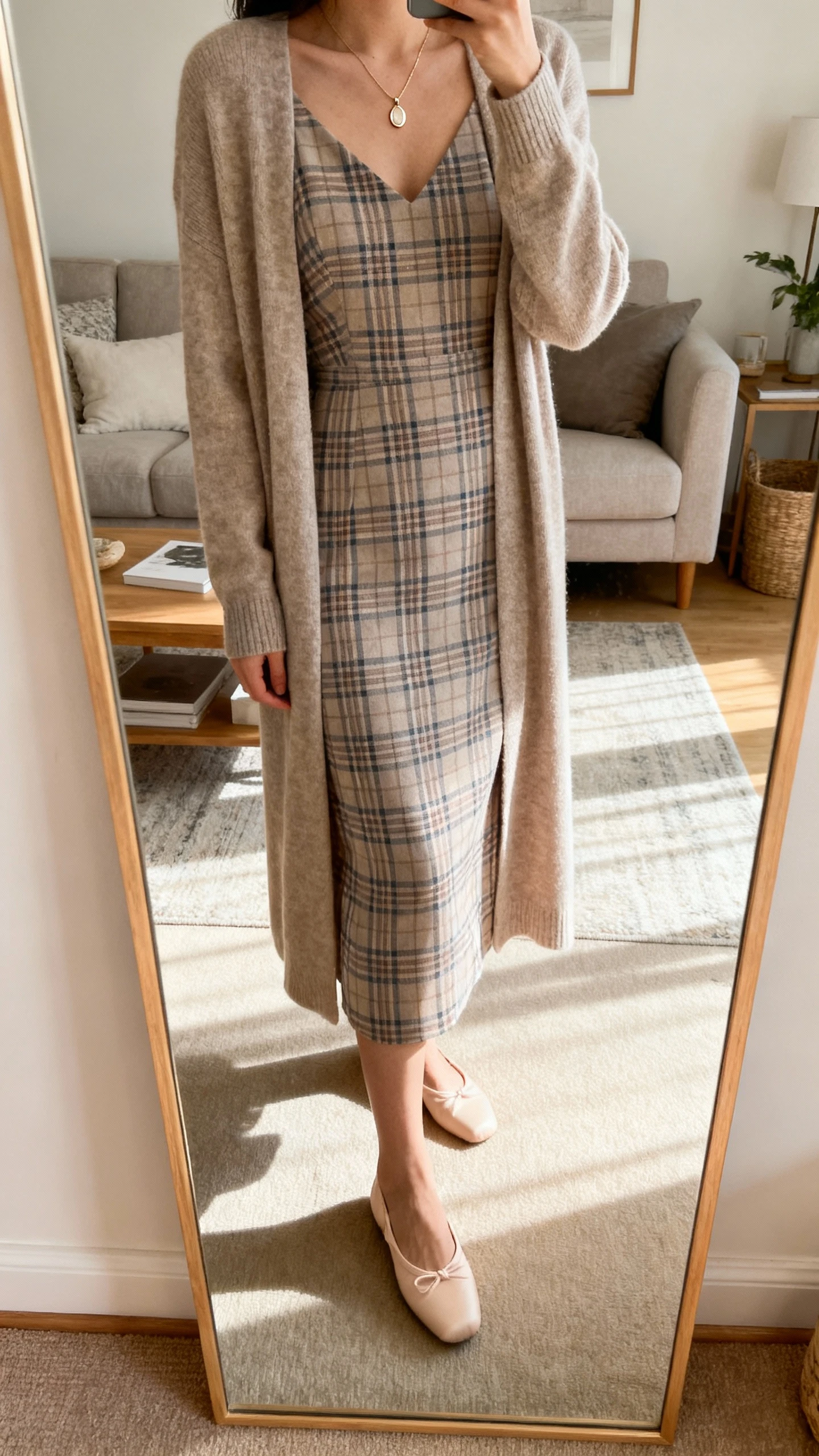iPhone mirror selfie of a woman wearing an elegant plaid dress under a longline cardigan, with ballet flats and a delicate pendant necklace; face not visible, cropped at shoulders, living room mirror, soft afternoon light, iPhone photo quality