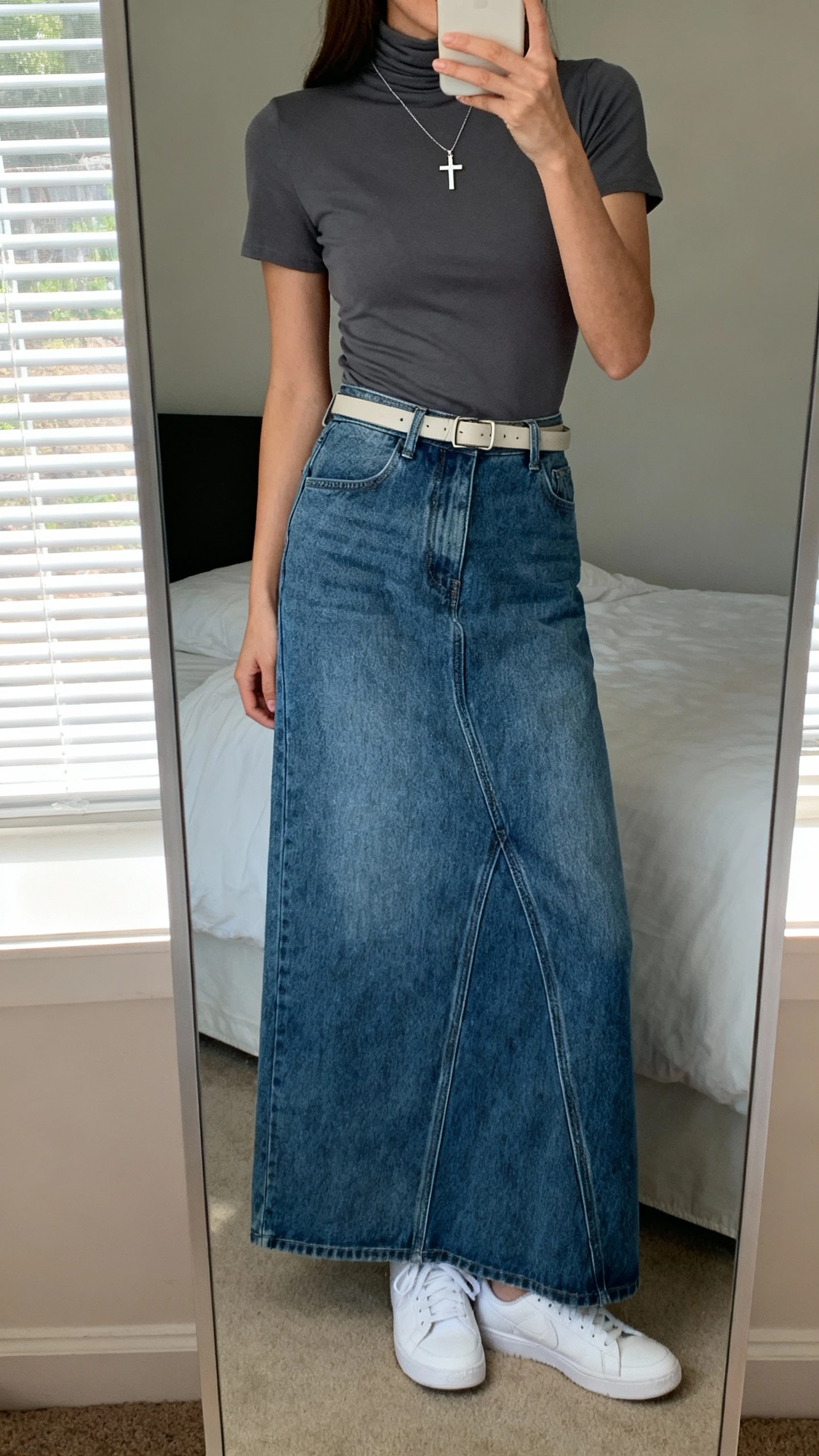 iPhone mirror selfie of a woman wearing an elevated denim maxi skirt, a fitted high-neck tee, minimalist belt, simple cross pendant necklace, and clean white sneakers, face not visible, cropped at neck, bedroom mirror, natural window light, iPhone photo quality.