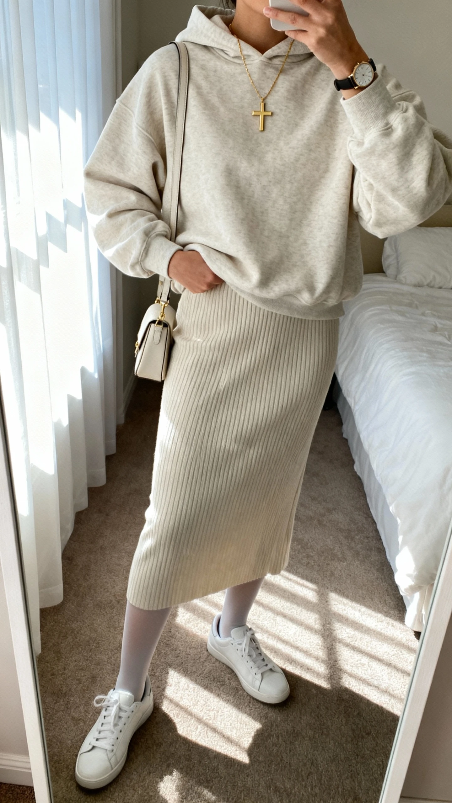 iPhone mirror selfie of a woman wearing an elevated oversized hoodie tucked into a ribbed midi skirt with opaque tights, low-profile white sneakers, delicate gold cross necklace, small shoulder bag, and minimalist watch; face not visible, cropped at neck, bedroom mirror, soft morning window light, iPhone photo quality.