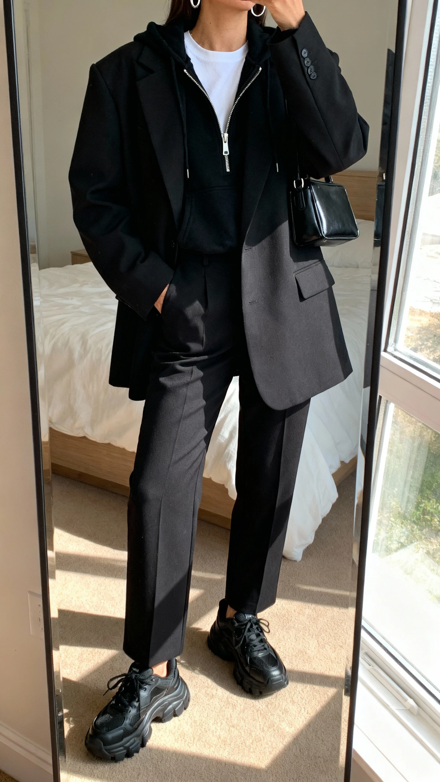 iPhone mirror selfie of a woman wearing an elevated monochrome set: oversized black blazer, matching black tailored trousers, crisp white tee layered under a black zip hoodie peeking out, chunky black dad sneakers, minimal silver hoops, black structured mini bag, face not visible, cropped at neck, bedroom mirror, natural window light, iPhone photo quality.