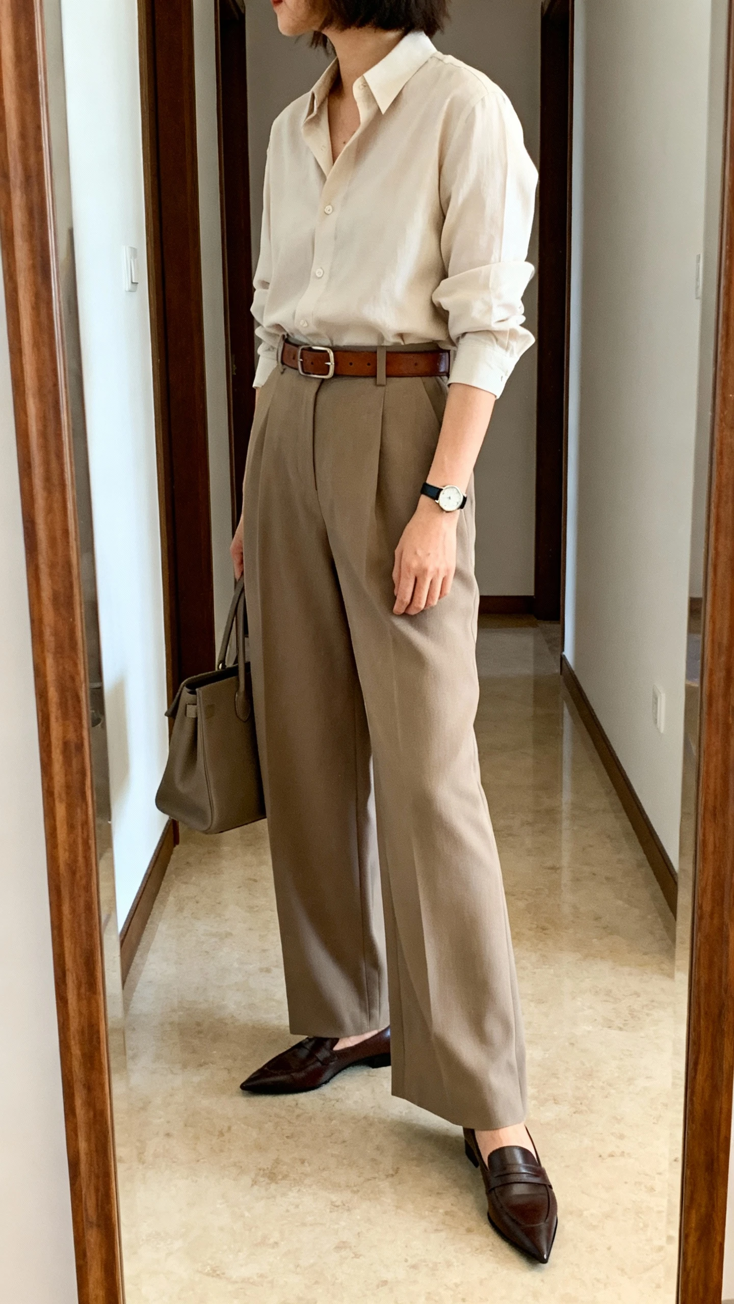 iPhone mirror selfie of a woman wearing an elevated blouse tucked into high-waisted wide-leg trousers, classic leather belt, pointed-toe loafers, slim watch, and a structured tote, face not visible, side angle in a hallway full-length mirror, morning indoor lighting, iPhone photo quality.