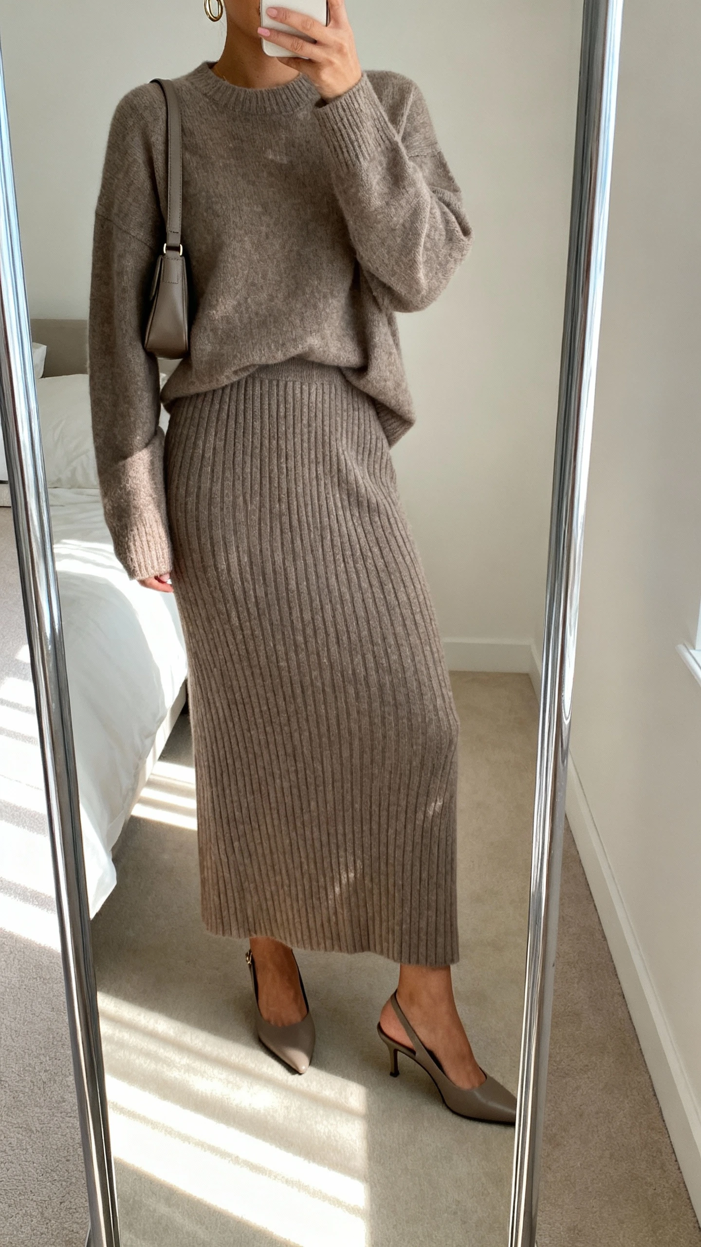 iPhone mirror selfie of a woman wearing an elevated monochrome knit set: long rib-knit skirt and matching knit sweater in taupe, sleek slingback heels, small shoulder bag, and subtle hoop earrings, face not visible, cropped at shoulders, bedroom mirror with soft morning light, iPhone photo quality