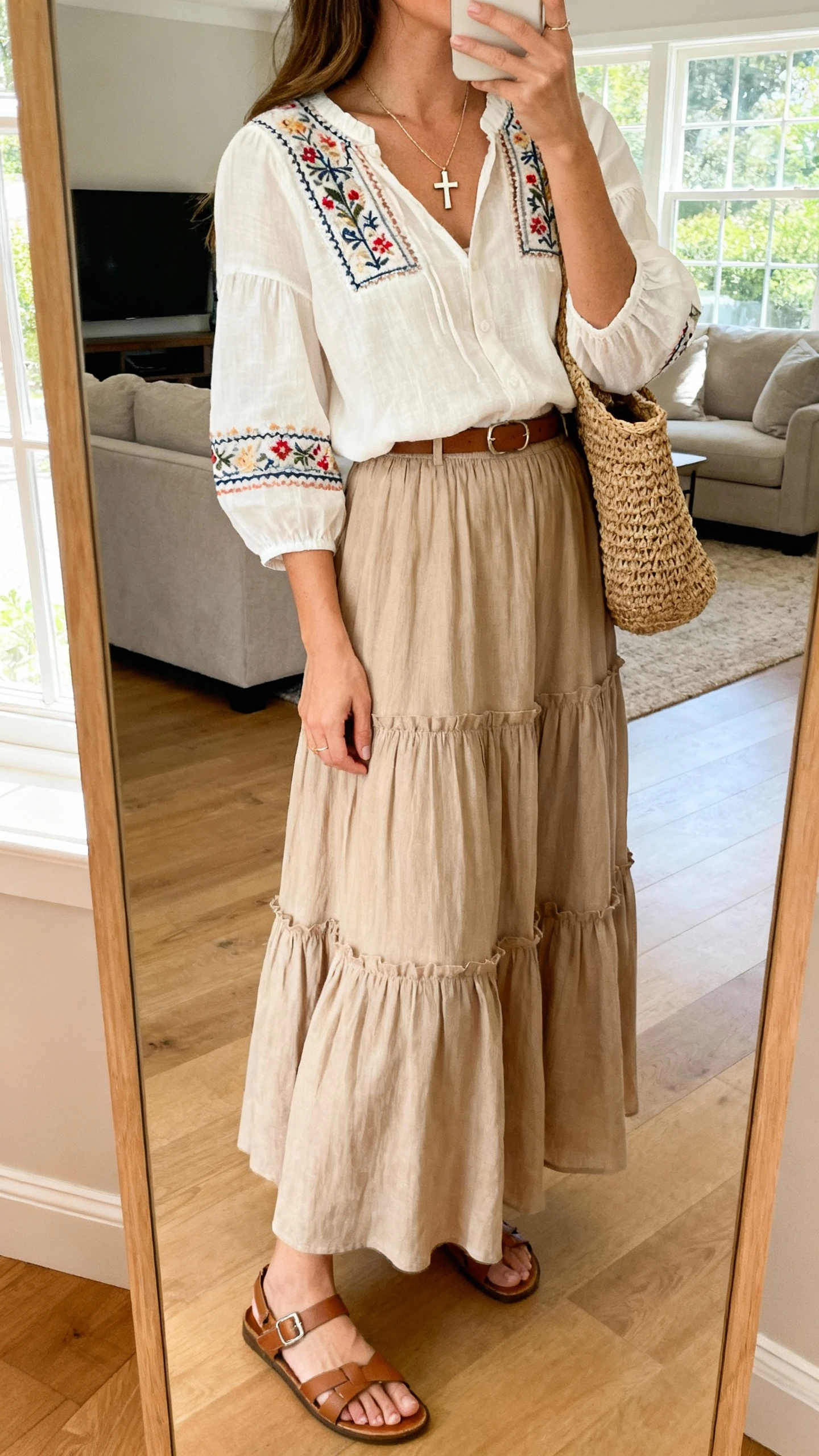 iPhone mirror selfie of a woman wearing an embroidered blouse tucked into a tiered maxi skirt, dainty cross necklace, woven tote, and sandals, face not visible, side angle, living room full-length mirror, bright natural window light, iPhone photo quality.