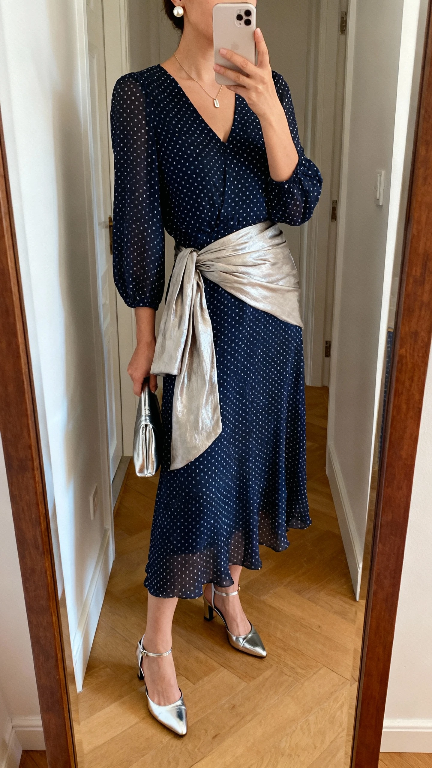 iPhone mirror selfie of a woman wearing an ink-blue polka dot chiffon midi dress with modest neckline and sleeves, lightweight soft metallic wrap, low-heel metallic slingbacks, pearl studs, slim pendant necklace, and a small metallic clutch, face not visible, cropped at neck, entryway full-length mirror, warm indoor lighting, iPhone photo quality.