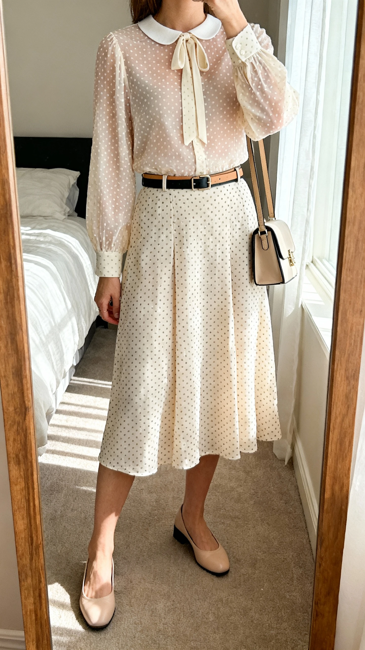 iPhone mirror selfie of a woman wearing an opaque Swiss dot blouse with a modest tie-neck/Peter Pan/crew neckline and a coordinating fully lined Swiss dot A-line midi skirt, thin leather belt in tan or black, classic ballet flats or low block heels, structured crossbody or small satchel; face not visible, cropped at shoulders, bedroom mirror with soft daylight, iPhone photo quality.