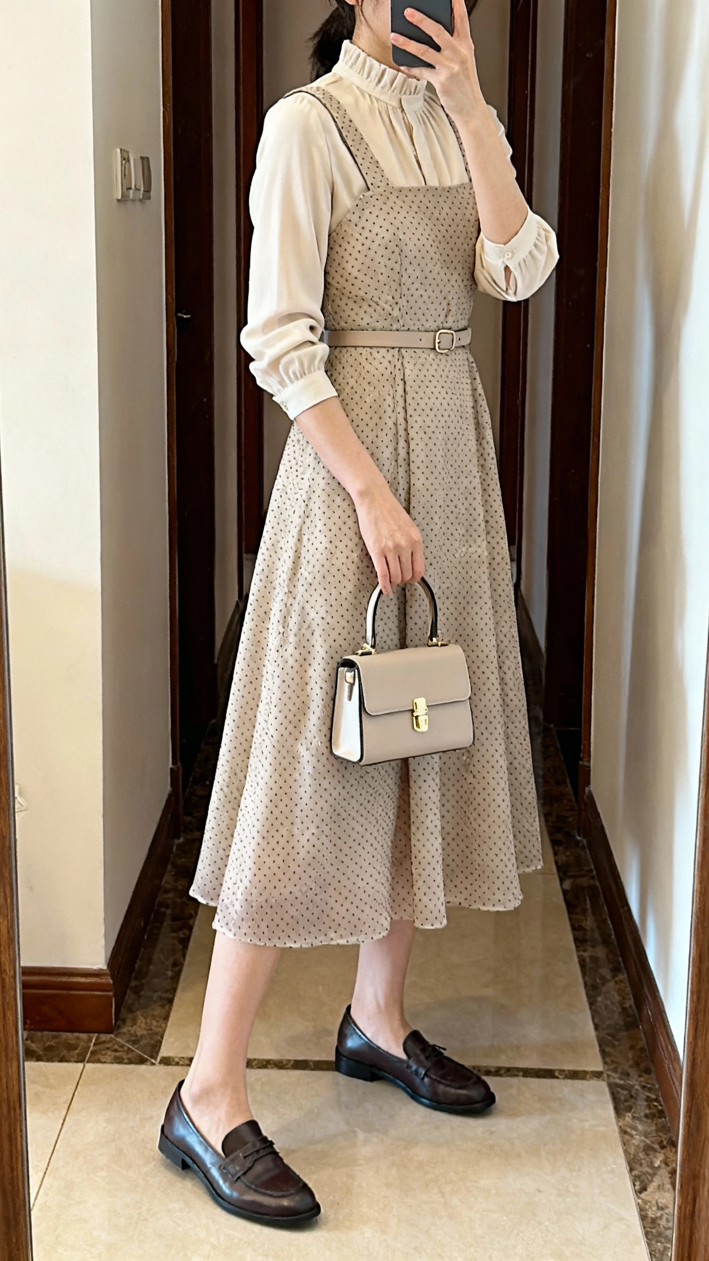 iPhone mirror selfie of a woman wearing an opaque Swiss dot pinafore midi with wide straps and modest A-line over a high-neck blouse (long or 3/4 sleeves), subtle waist belt or built-in tie, closed-toe Oxford flats/Mary Janes/ankle-strap block heels, compact top-handle bag; face not visible, side angle in entryway full-length mirror, warm indoor lighting, iPhone photo quality.