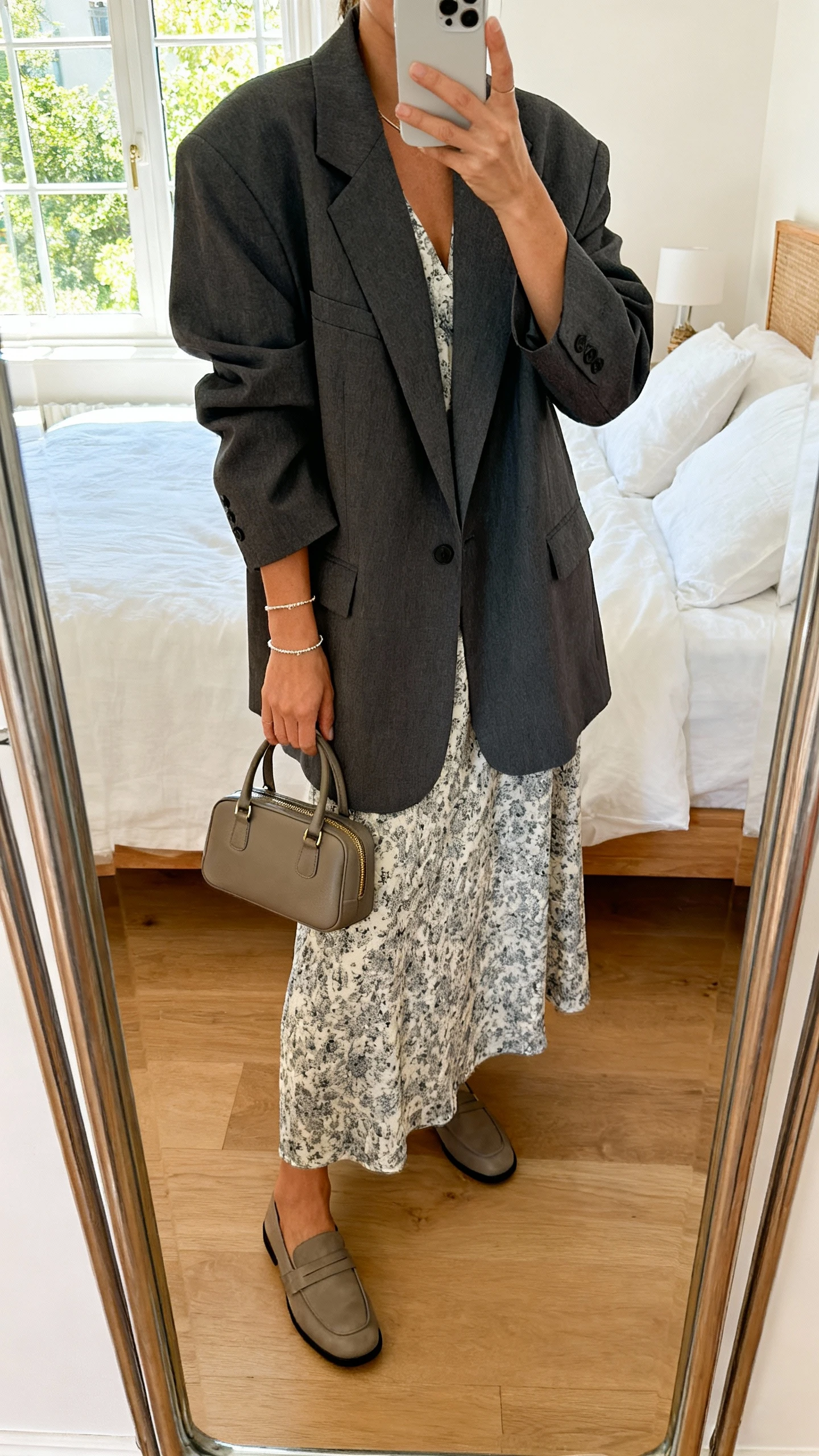 iPhone mirror selfie of a woman wearing an oversized charcoal blazer over a printed maxi dress with loafers, structured mini handbag, delicate bracelet, face not visible, cropped at shoulders, bedroom mirror, bright natural window light, iPhone photo quality.