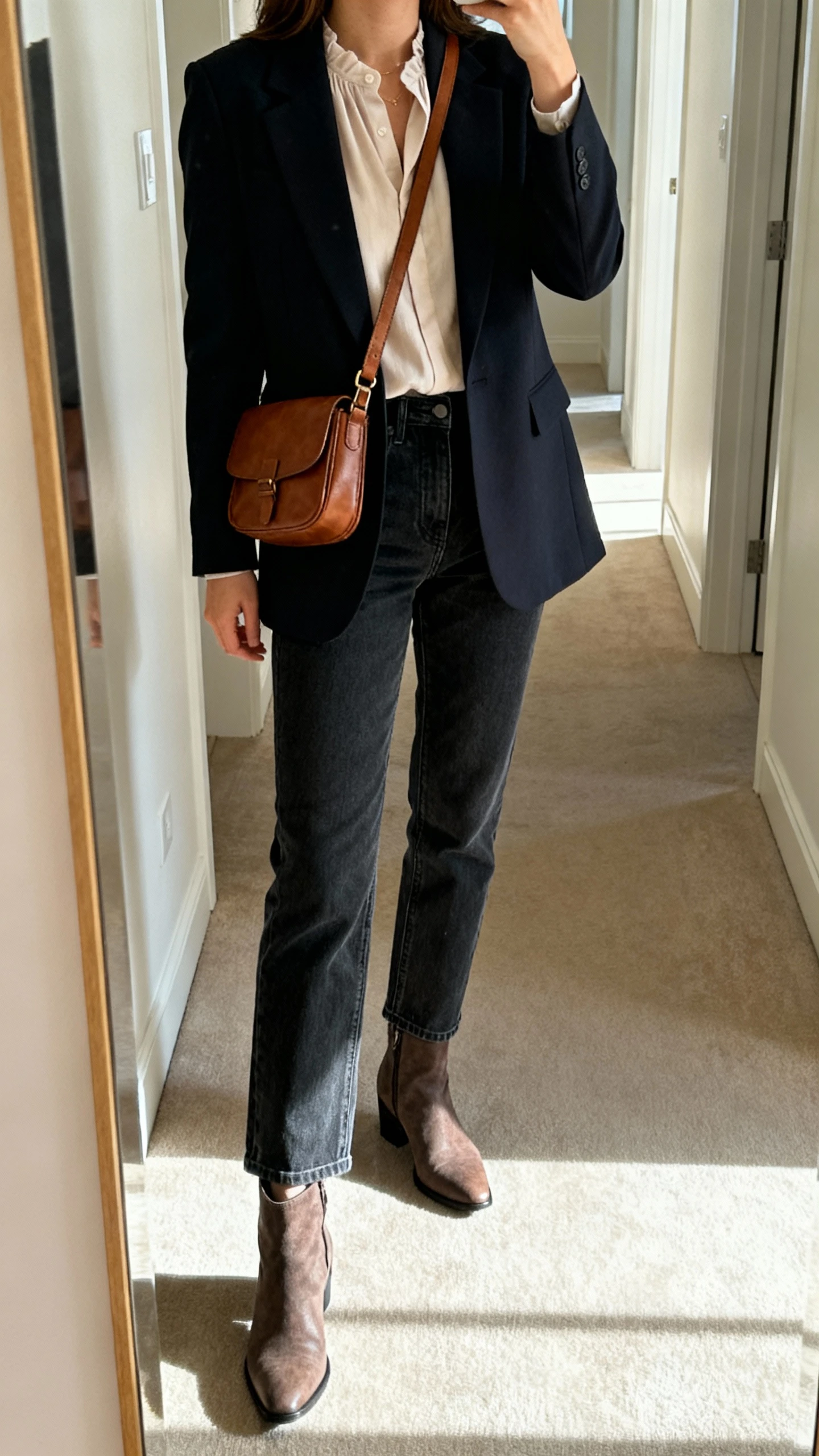 iPhone mirror selfie of a woman wearing dark-wash straight jeans, tailored blazer, soft blouse, ankle boots, and a leather crossbody, face not visible, cropped at shoulders, full-length hallway mirror with indoor daylight, iPhone photo quality.
