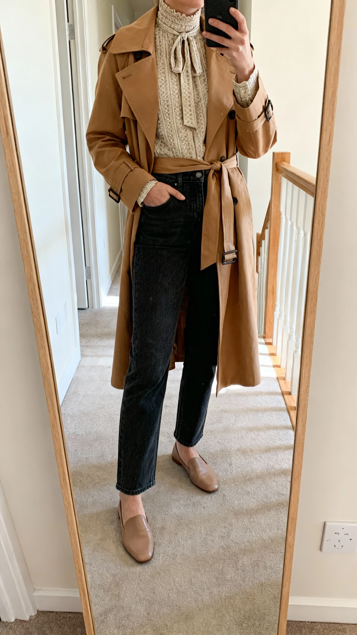 iPhone mirror selfie of a woman wearing full-length straight dark-wash jeans, a high-neck woven blouse with a tie-neck, a camel belted trench coat, and soft leather flats; face not visible, full-length hallway mirror, natural daylight, iPhone photo quality.
