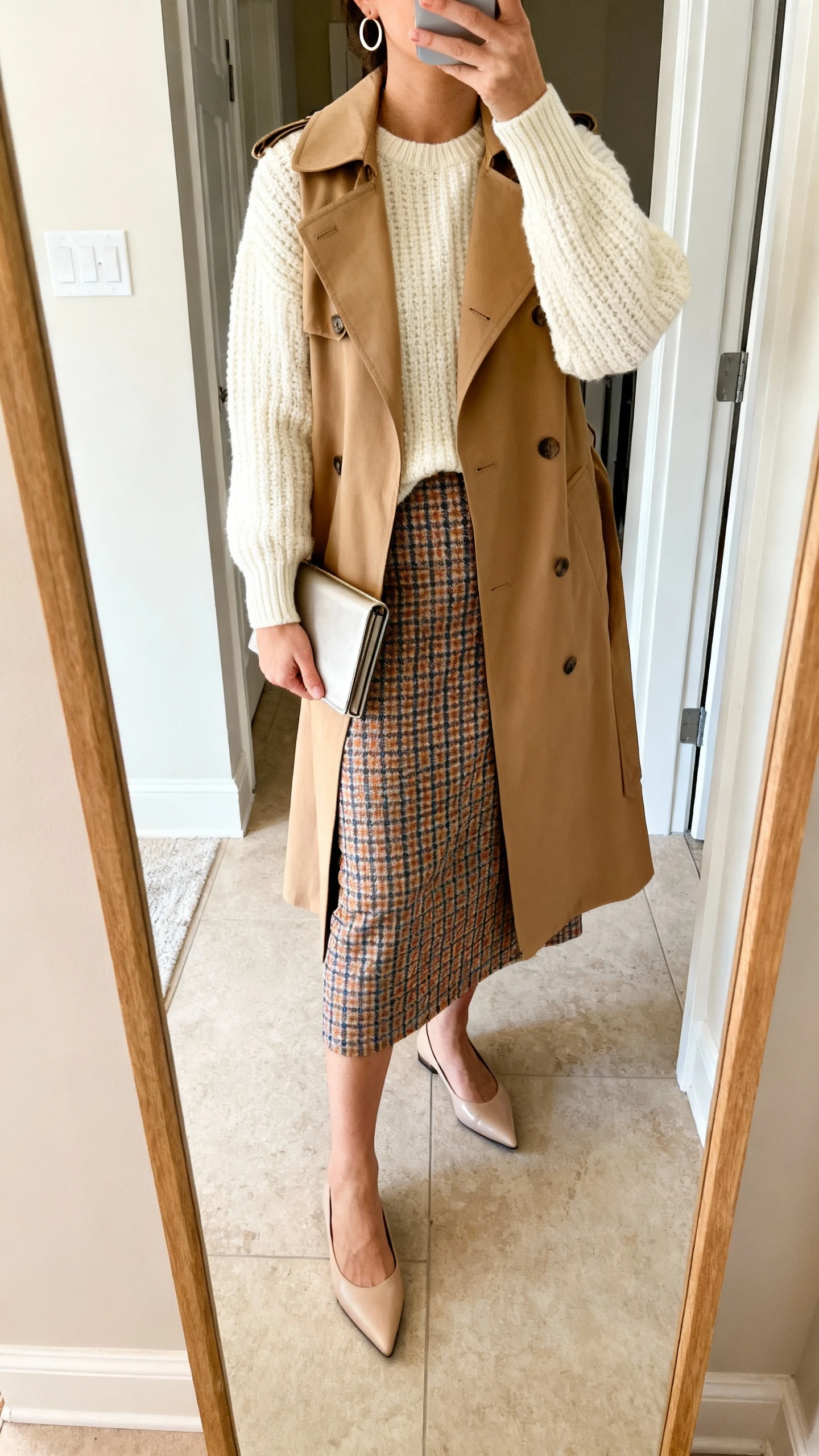 iPhone mirror selfie of a woman wearing monochrome layers (cream knit and tan trench) with a plaid skirt as the pop, pointed flats, minimal hoop earrings, and a slim clutch, face not visible, cropped at neck, entryway full-length mirror, soft indoor lighting, iPhone photo quality.