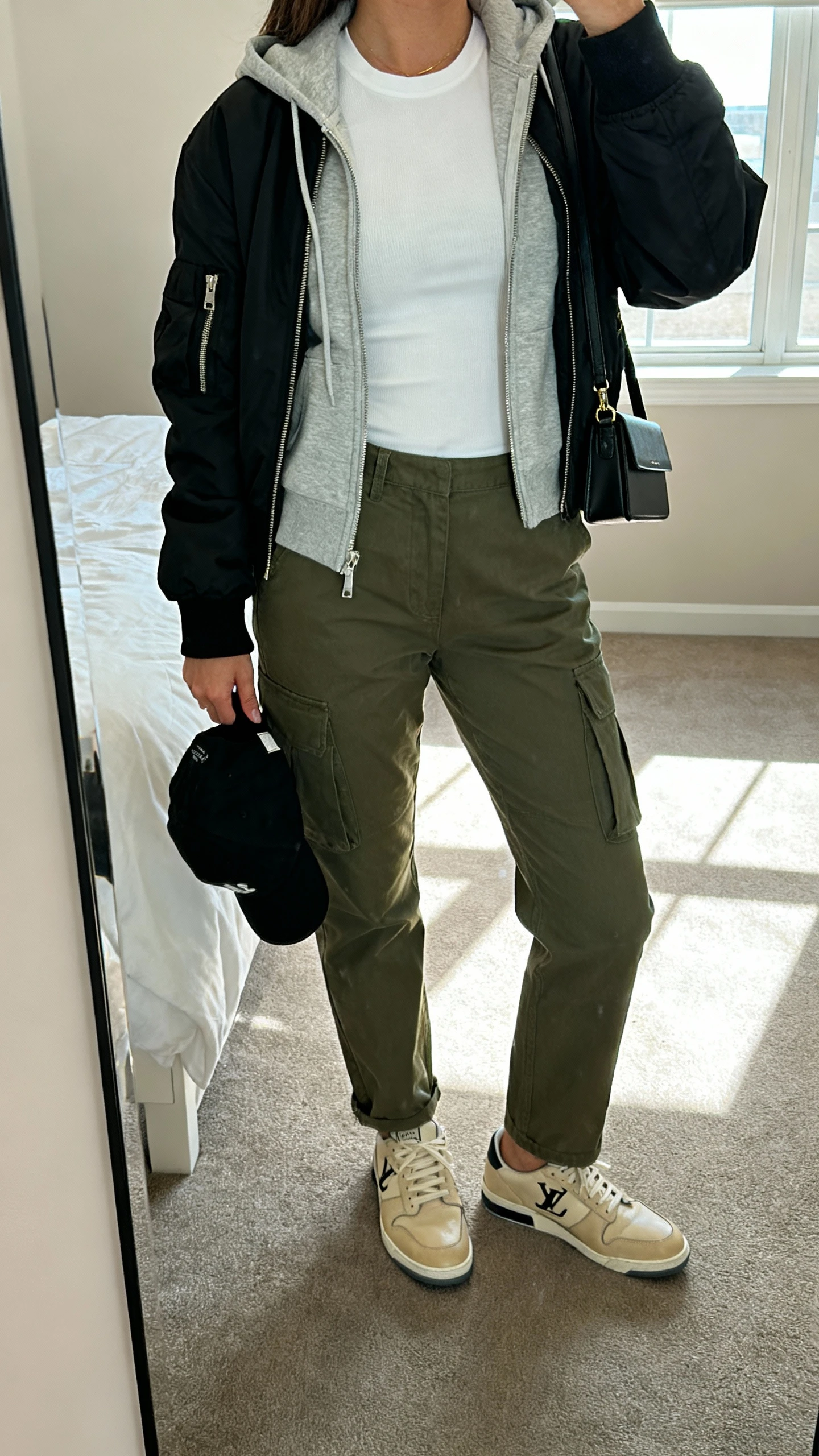 iPhone mirror selfie of a woman wearing olive cargo pants, white fitted tee, light grey zip-up hoodie layered under a black bomber, beige “quiet luxury” leather sneakers, black cap in hand, slim black crossbody, face not visible, cropped at shoulders, bedroom mirror, window light, iPhone photo quality.