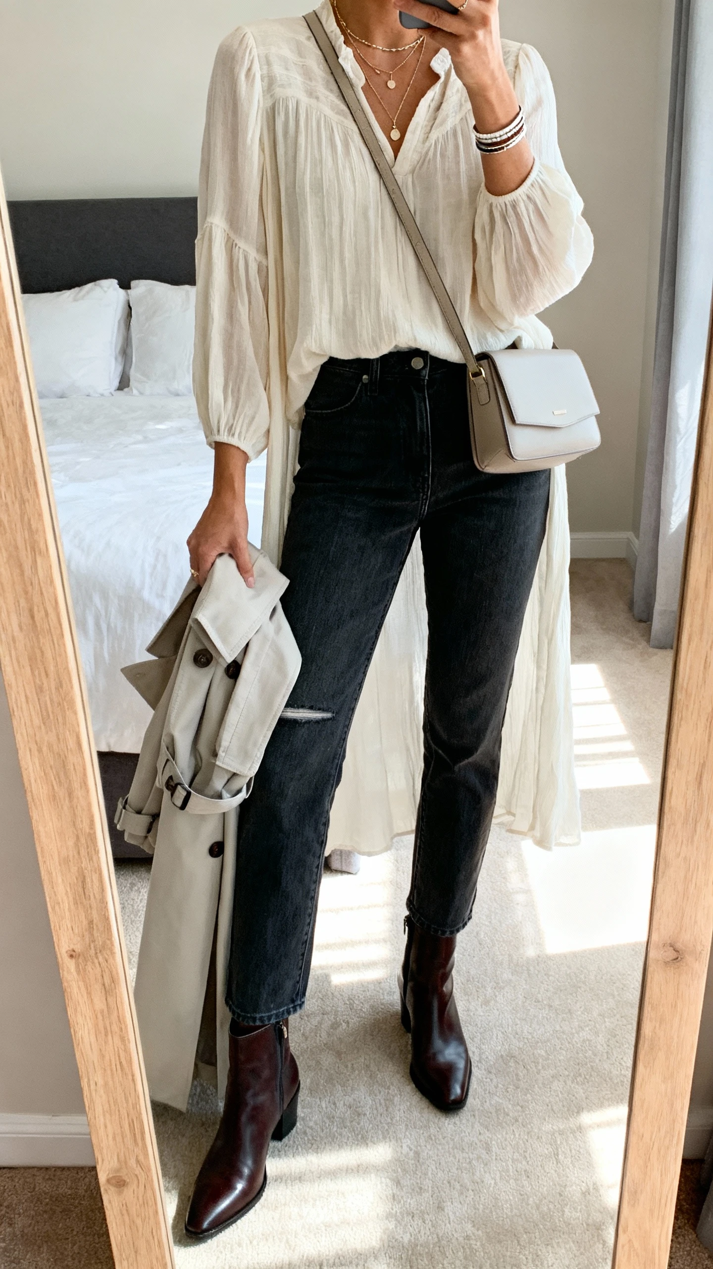 iPhone mirror selfie of a woman wearing straight-leg dark-wash jeans with no distressing, a flowy longline blouse, polished ankle boots, layered delicate necklaces with a simple bracelet stack, a minimal crossbody bag, and a light trench draped over the arm, face not visible, cropped at neck, bedroom full-length mirror, natural afternoon light, iPhone photo quality.