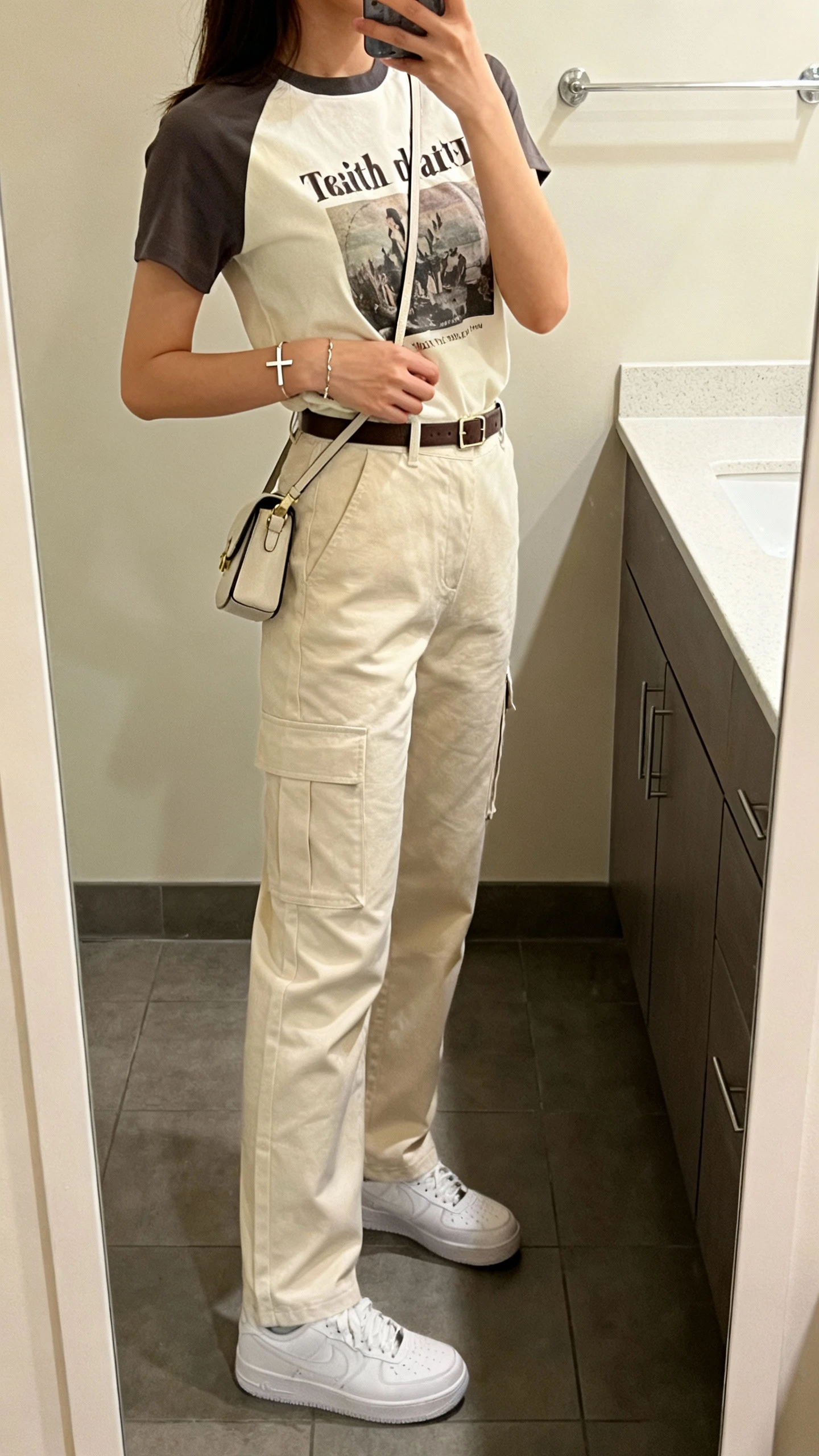 iPhone mirror selfie side angle of a woman wearing clean-cut cargo pants with a faith-focused graphic tee tucked in, slim belt, simple cross bracelet, white sneakers, small crossbody bag, face not visible, bathroom mirror, indoor soft lighting, iPhone photo quality.