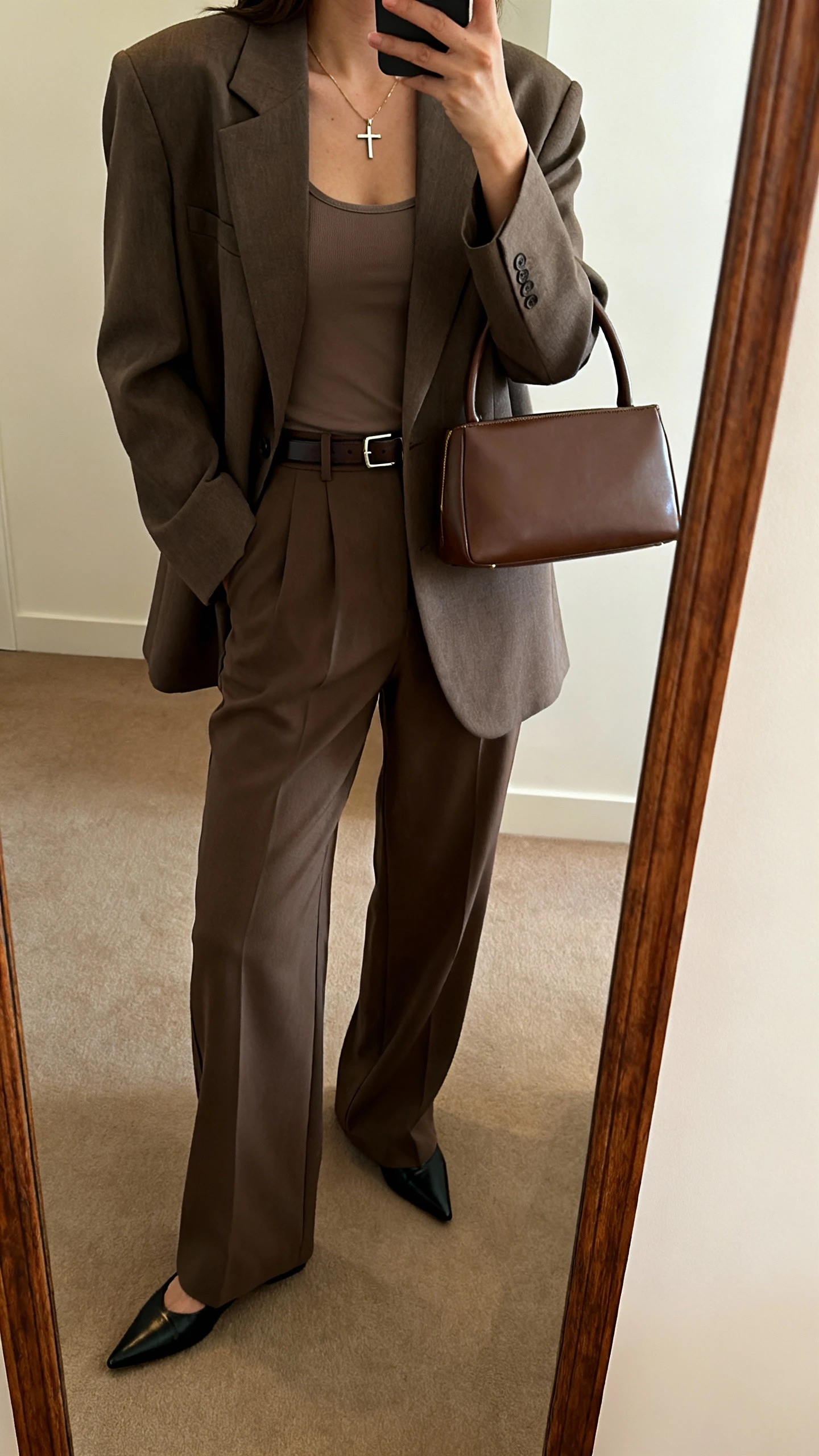 iPhone mirror selfie side angle of a woman wearing a boxy blazer with wide-leg trousers, modest camisole underneath, pointed flats, slim belt, dainty cross necklace, top-handle bag, face not visible, full-length mirror, indoor warm lighting, iPhone photo quality.