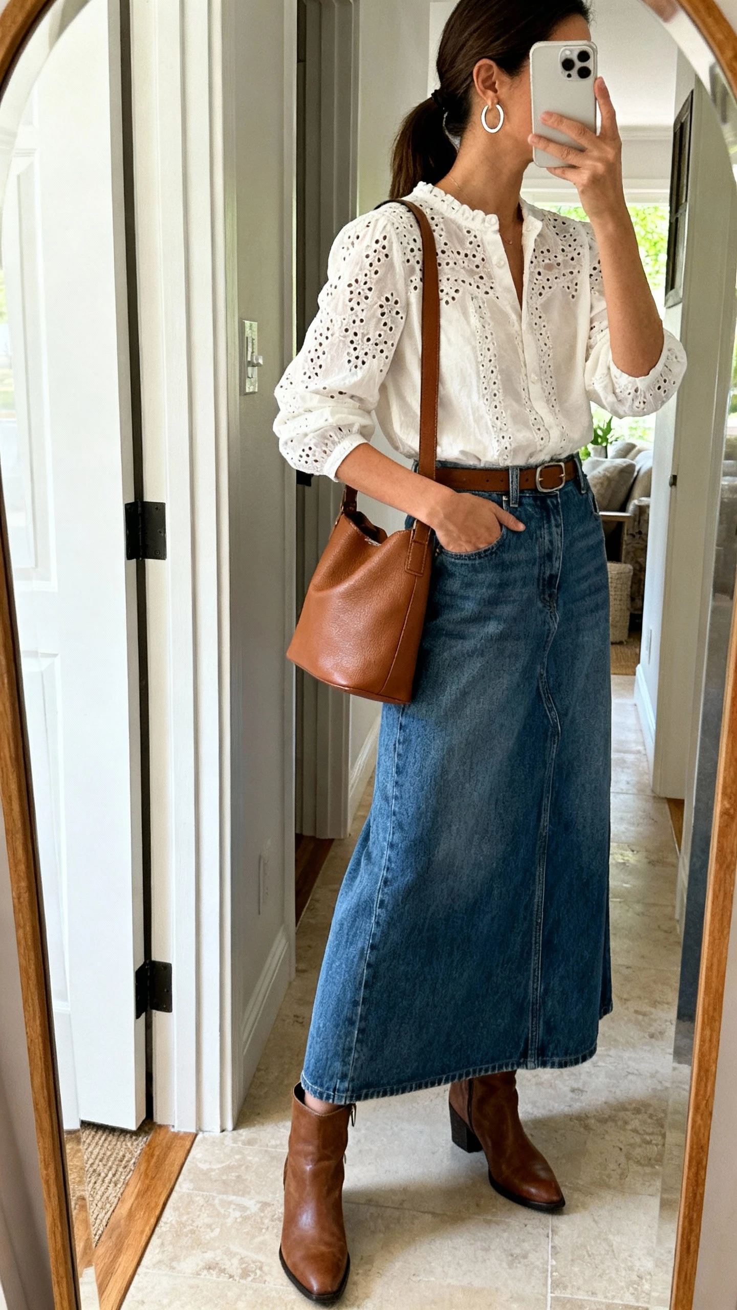 iPhone mirror selfie side profile of a woman wearing an eyelet detail blouse tucked into a denim maxi skirt, simple hoop earrings, leather shoulder bag, and ankle boots, face not visible, entryway mirror, natural indoor light, iPhone photo quality.