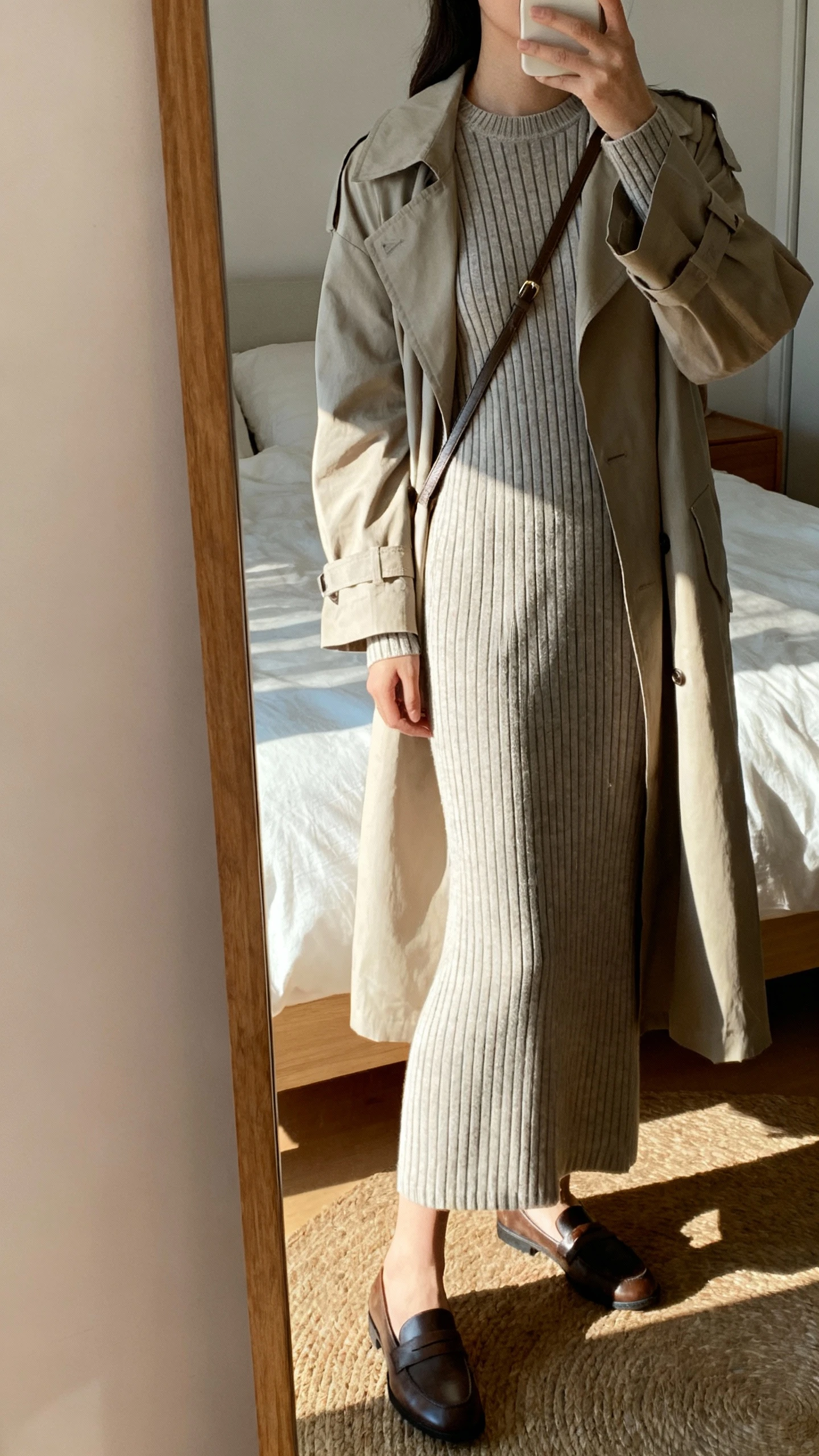 iPhone side-angle mirror selfie of a woman in a knit column dress with fine vertical stripes (ankle-grazing, crew neck, long sleeves), layered with a lightweight trench, paired with loafers and a slim crossbody; face not visible, cropped at shoulders, bedroom mirror, soft afternoon light, iPhone photo quality.