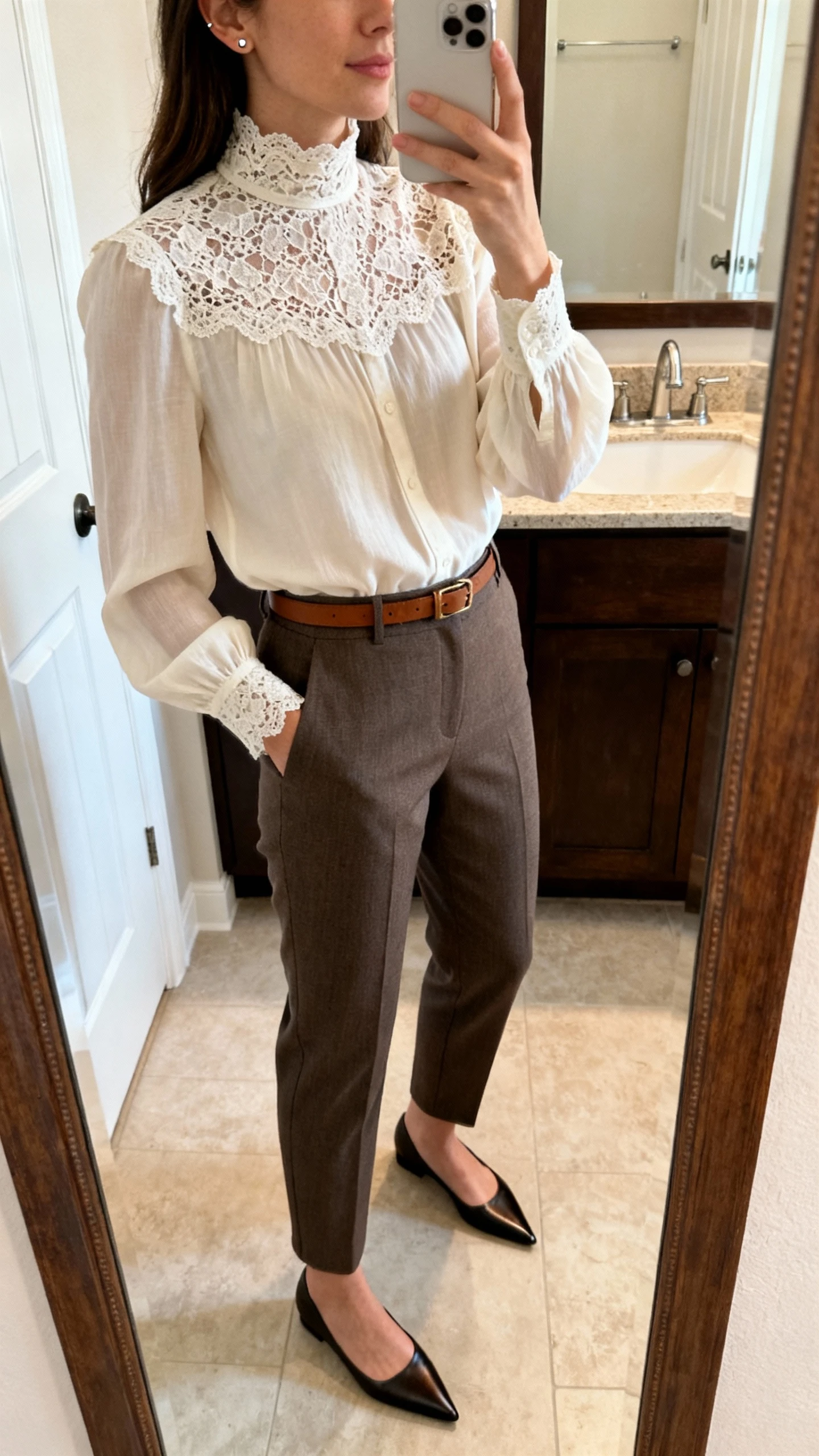 iPhone side-angle mirror selfie of a woman in a lace-trimmed high-neck blouse paired with tailored trousers, pointed flats, minimal stud earrings, and a slim leather belt, face not visible, bathroom mirror, soft morning light, iPhone photo quality.