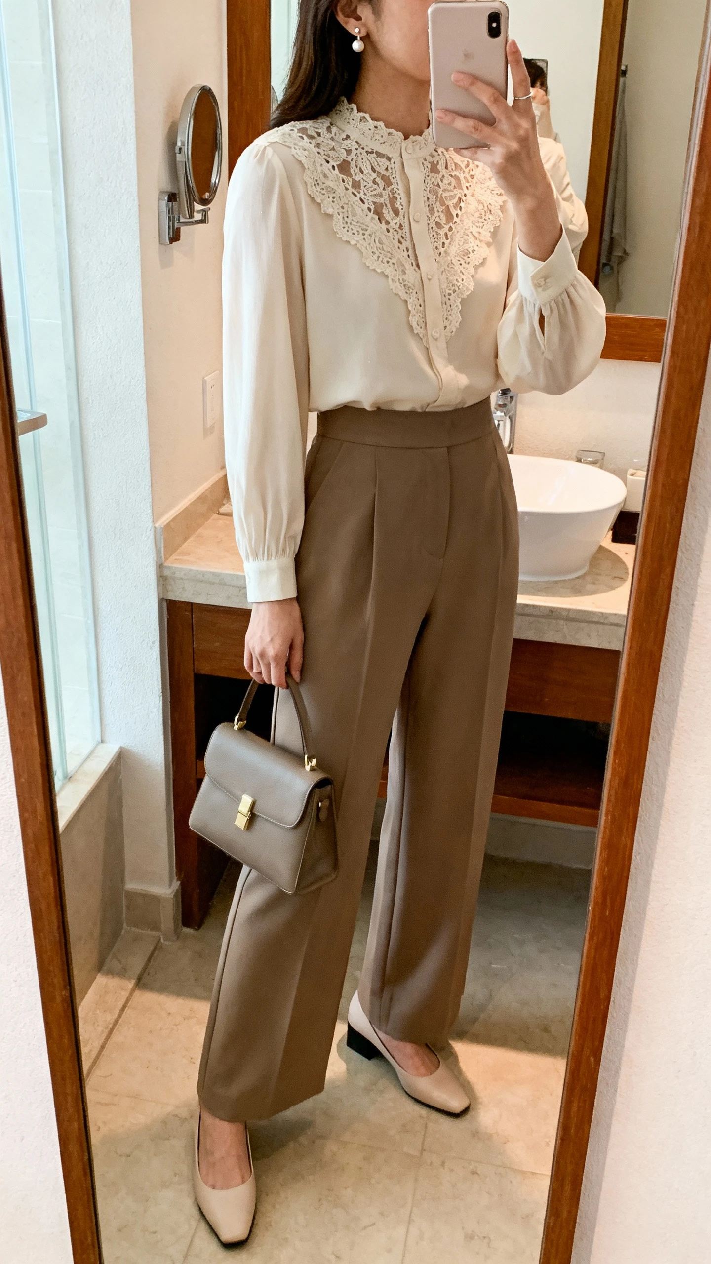 iPhone side-angle mirror selfie of a woman in a lace-trim blouse with high-waisted wide-leg trousers, delicate stud earrings, low block heels, and a structured handbag, face not visible, bathroom mirror, natural indoor light, iPhone photo quality.