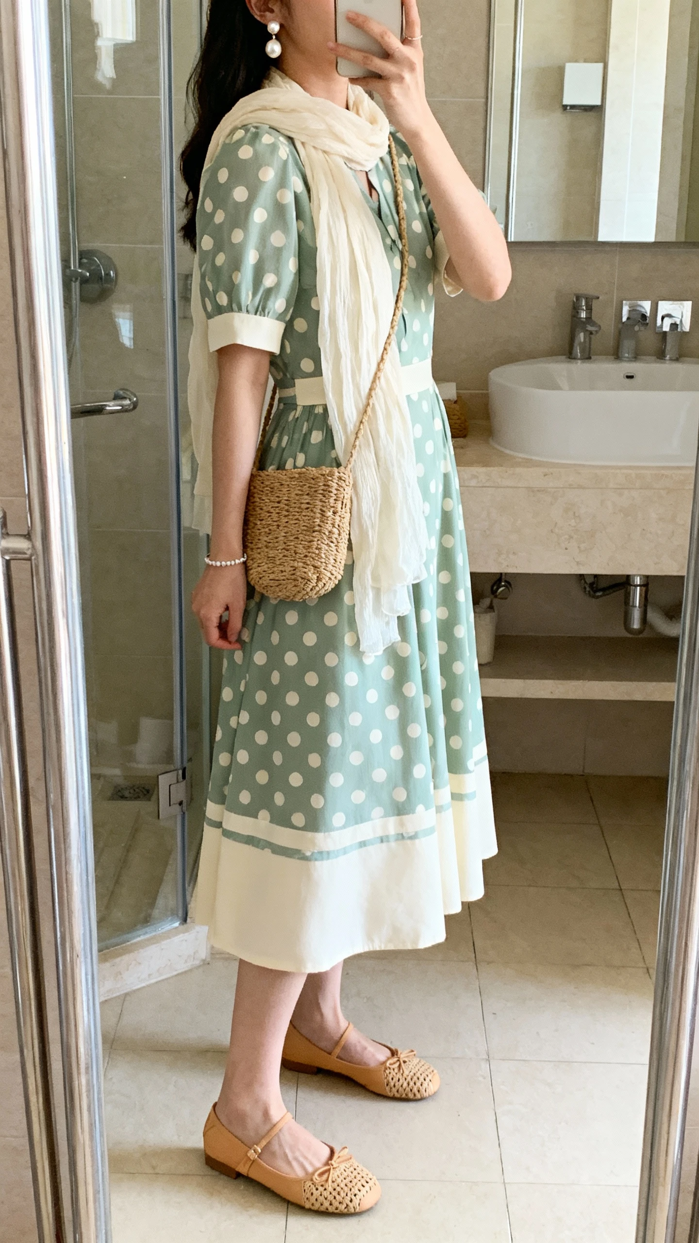 iPhone side-angle mirror selfie of a woman in a pastel polka dot tea-length dress (sage-and-cream), lightweight cream chiffon scarf, woven tan ballet flats, pearl drop earrings, dainty bracelet, and a straw crossbody, face not visible, bathroom full-length mirror, morning light, iPhone photo quality.