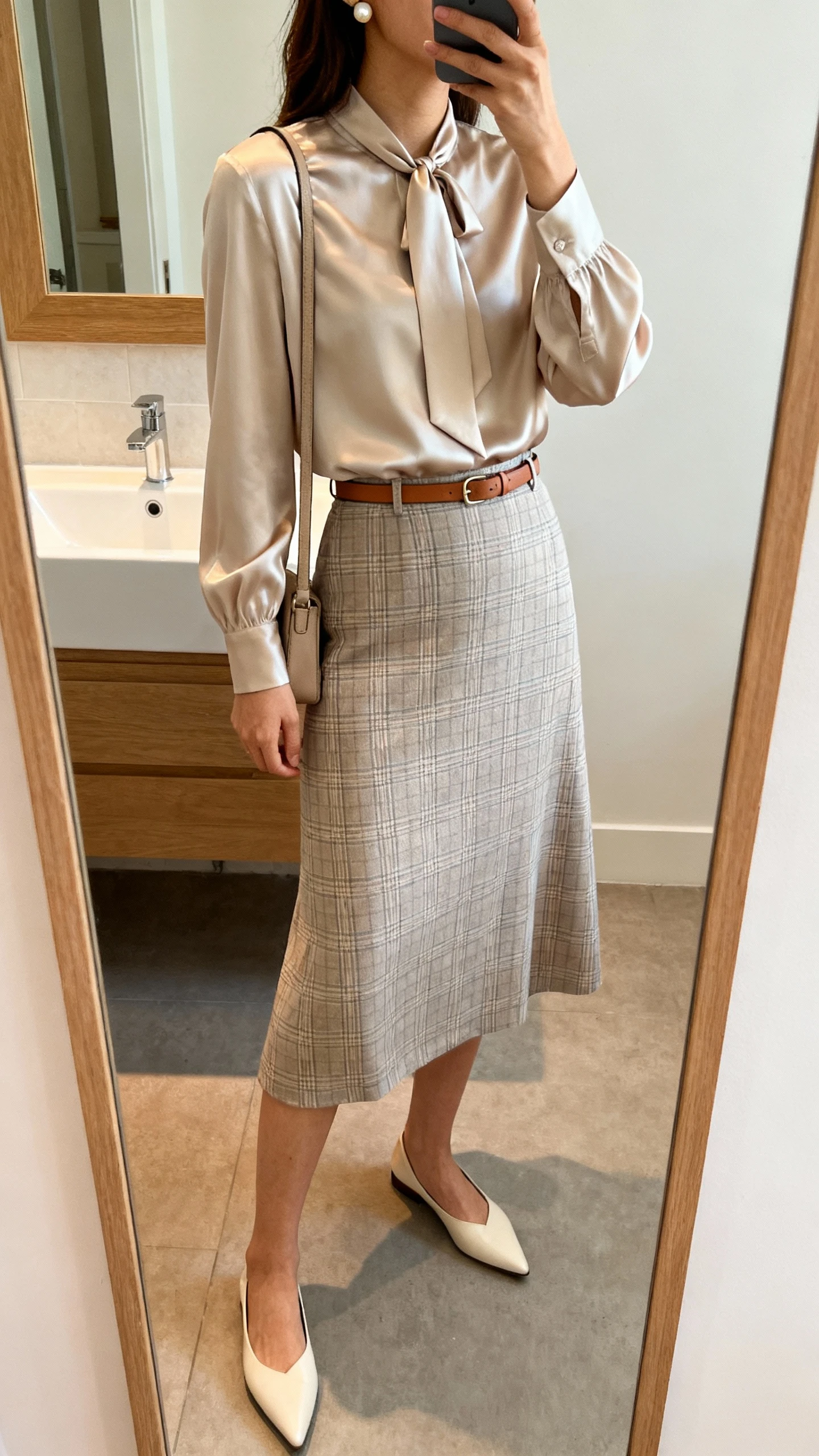 iPhone side-angle mirror selfie of a woman in a silky tie-neck blouse tucked into an A-line midi skirt (subtle plaid), pointed flats, a thin leather belt, a minimal crossbody bag, and pearl stud earrings; face not visible, bathroom mirror, soft indoor lighting, iPhone photo quality.