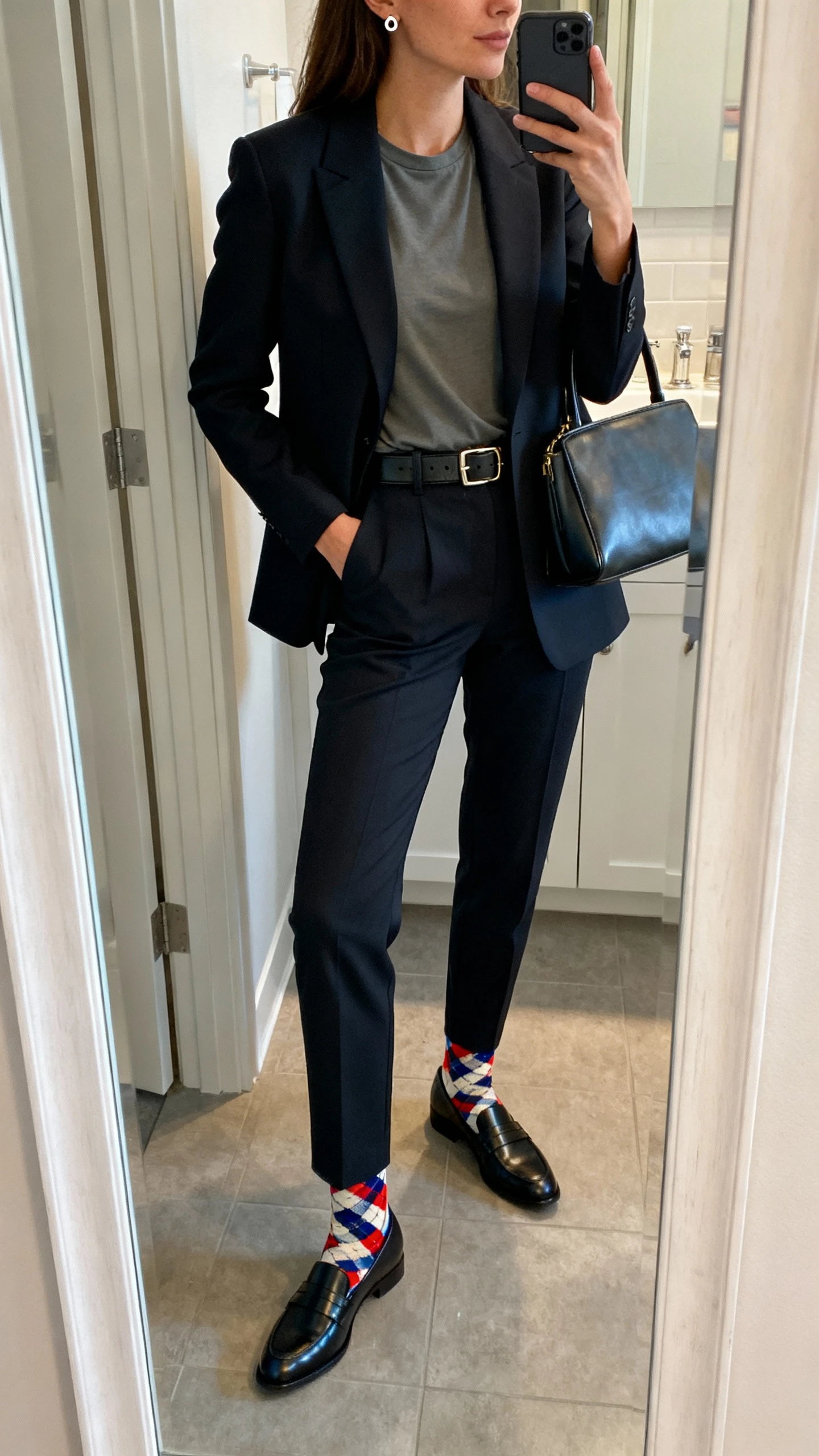 iPhone side-angle mirror selfie of a woman in a tailored blazer over a modest crew tee, straight-leg trousers, sleek leather belt, loafers, playful patterned socks, structured tote, and subtle stud earrings; face not visible, cropped at shoulders, bathroom mirror, indoor lighting, iPhone photo quality.