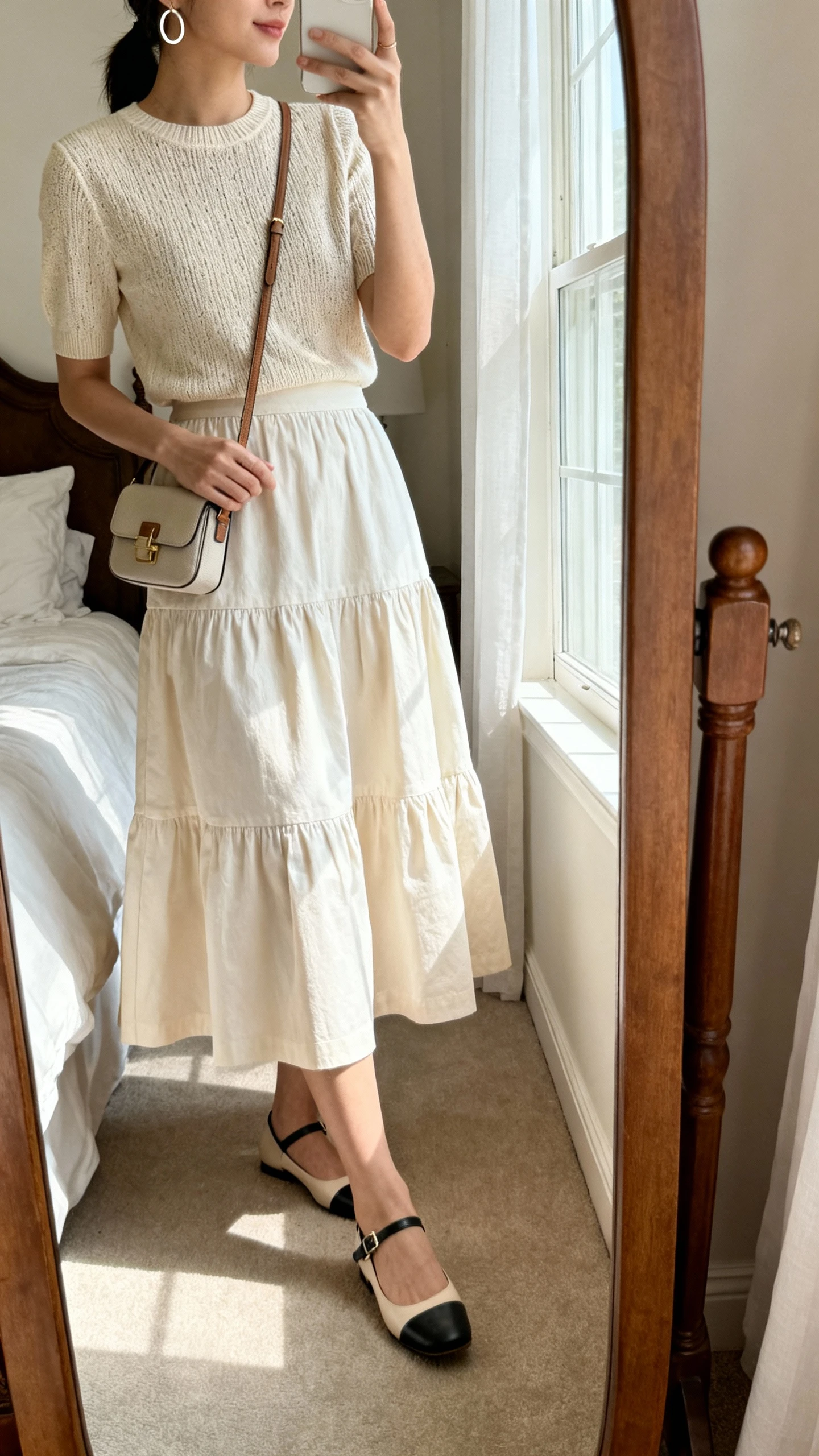 iPhone side-angle mirror selfie of a woman in a tiered cotton midi skirt paired with a polished short-sleeve knit top, ankle-strap flats, a petite crossbody, and simple hoop earrings, face not visible, cropped at shoulders, bedroom mirror, soft window light, iPhone photo quality.