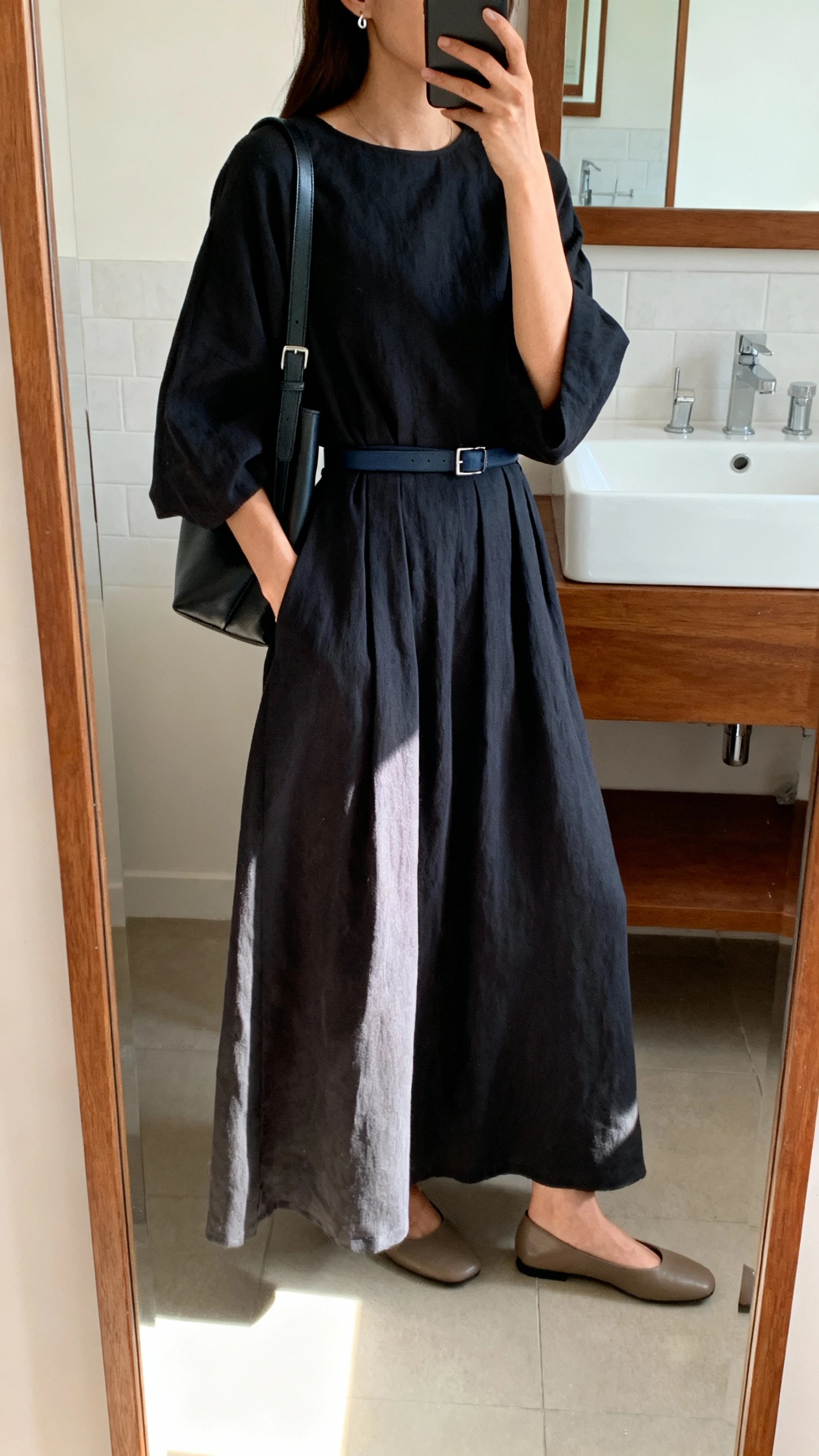 iPhone side-angle mirror selfie of a woman wearing an elevated monochrome loose-fit maxi dress in deep charcoal with a matching slim belt, structured shoulder bag, minimalist leather flats, and subtle stud earrings, face not visible, cropped at shoulders, bathroom full-length mirror, indoor natural light, iPhone photo quality