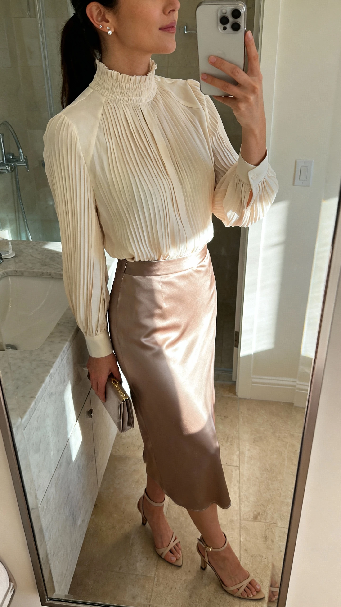 iPhone side-angle mirror selfie of a woman wearing a soft-pleat high-neck blouse tucked into a satin midi skirt, delicate stud earrings, ankle-strap heels, and a small clutch, face not visible, bathroom mirror with morning light, iPhone photo quality.