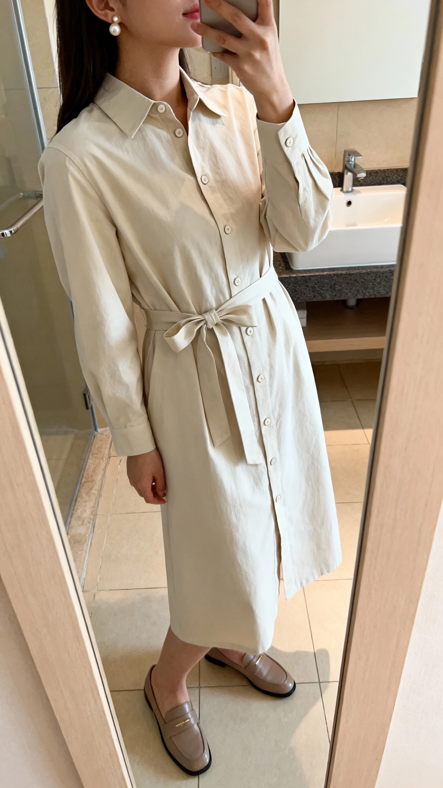 iPhone side-angle mirror selfie of a woman wearing a classic cotton shirt dress with a button front, collared neckline, and a belted bow at the waist, styled with simple pearl studs and loafers, face not visible, bathroom mirror, soft indoor lighting, iPhone photo quality