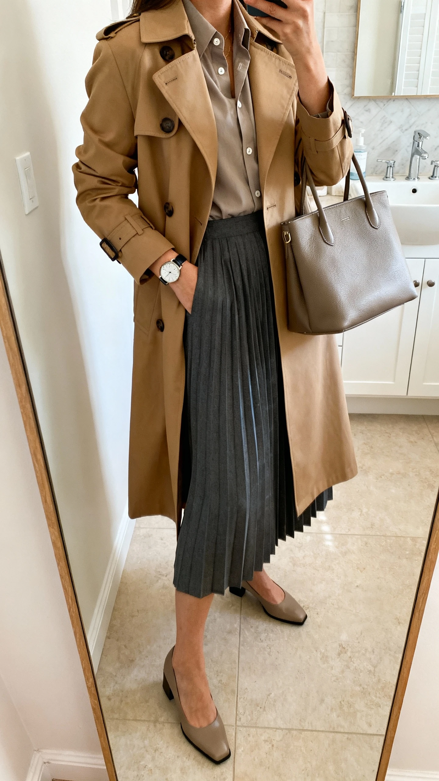 iPhone side-angle mirror selfie of a woman wearing a tailored camel trench coat over a crisp button-up and a midi pleated skirt in charcoal, structured tote bag, classic watch, and low block heels, face not visible, bathroom full-length mirror, morning indoor lighting, iPhone photo quality