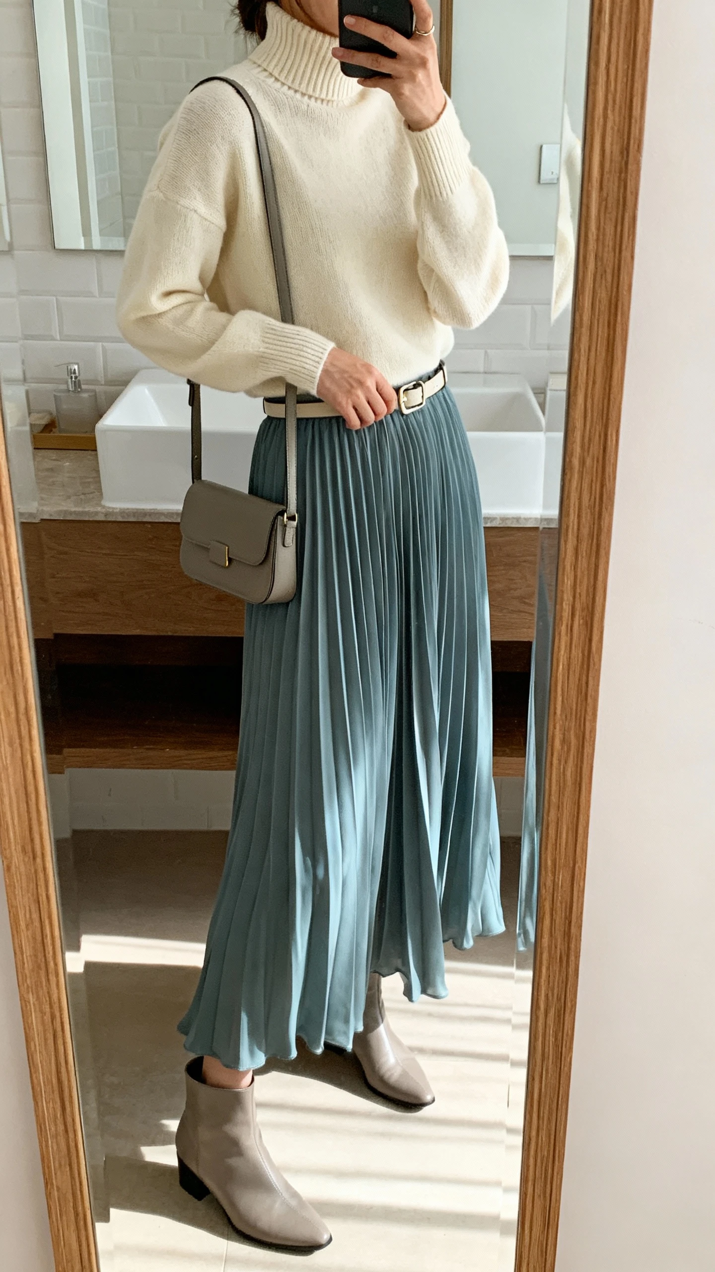iPhone side-angle mirror selfie of a woman wearing a soft cream mock-neck knit with a flowy dusty blue pleated maxi skirt, elegant ankle boots, slim waist belt, and a structured crossbody bag, face not visible, bathroom mirror, morning light, iPhone photo quality.
