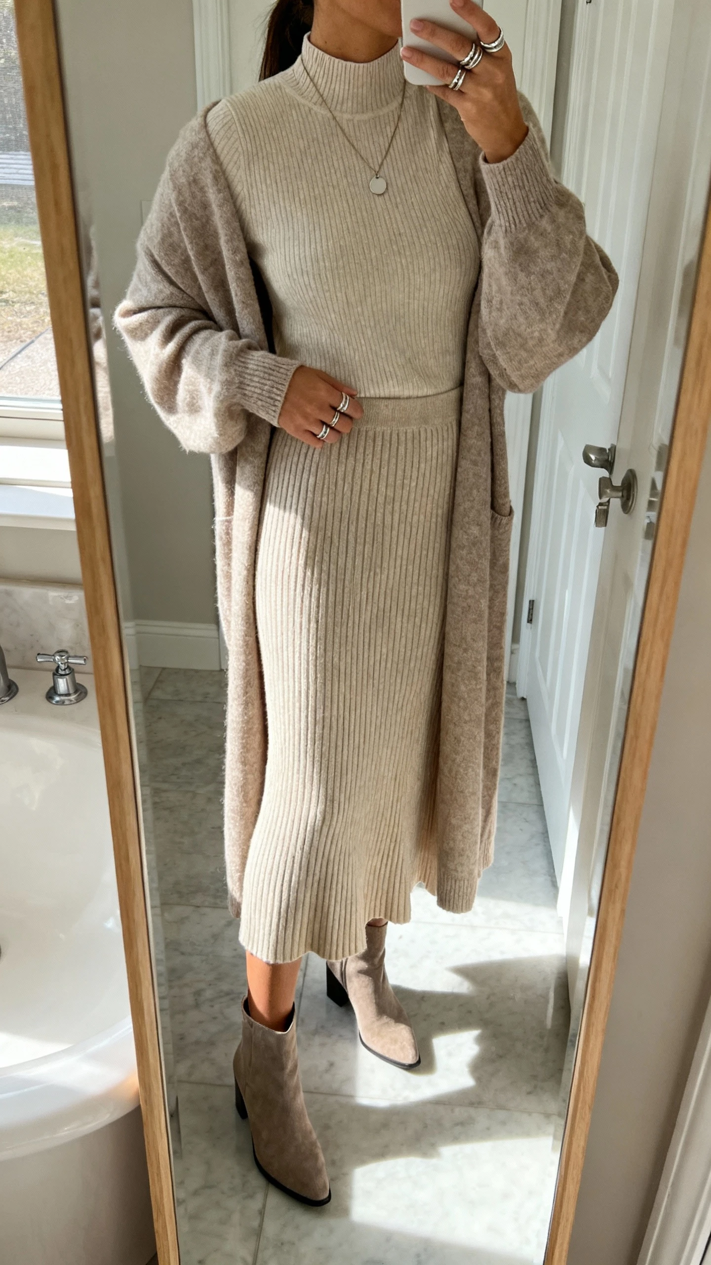 iPhone side-angle mirror selfie of a woman wearing a full-length ribbed knit skirt (slight A-line), a fine-gauge modest mock-neck knit top, a longline duster cardigan, low-heel ankle boots, a simple pendant necklace, and stackable rings, face not visible, bathroom mirror, soft indoor lighting, iPhone photo quality.
