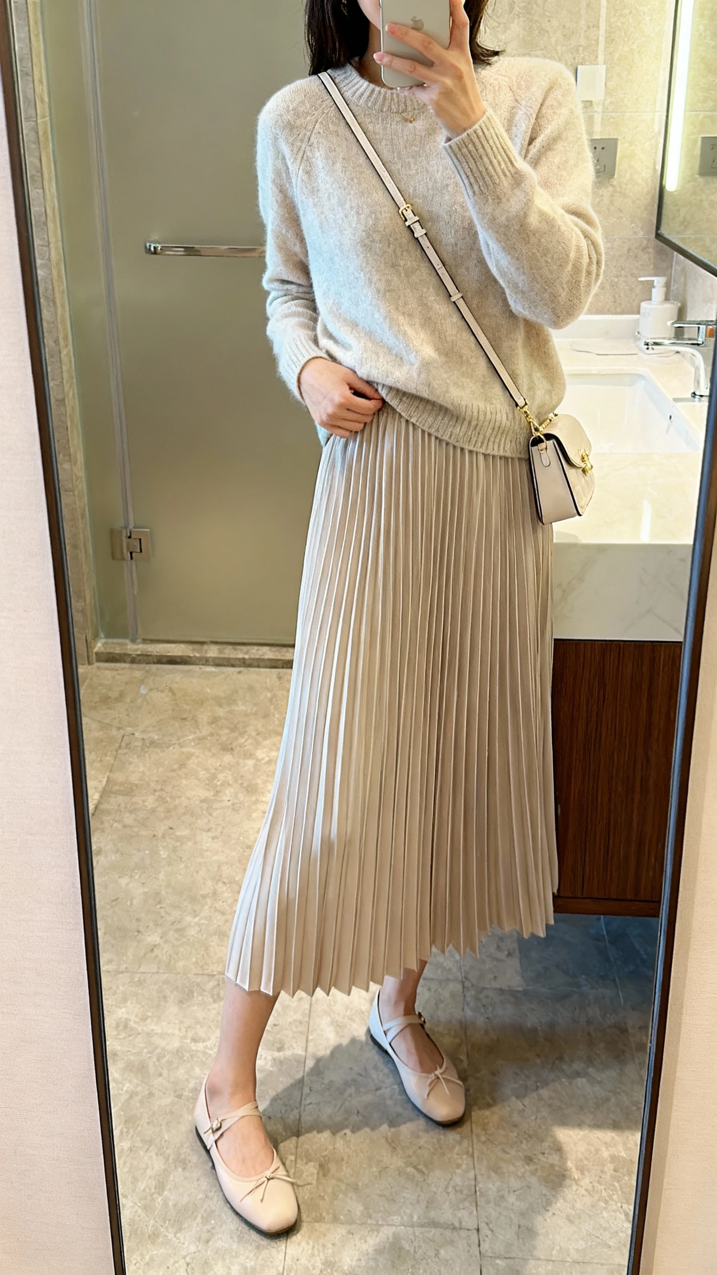 iPhone side-angle mirror selfie of a woman wearing a pleated midi skirt and a soft knit sweater tucked in, with ballet flats and a delicate crossbody bag, face not visible, bathroom full-length mirror, natural indoor lighting, iPhone photo quality.