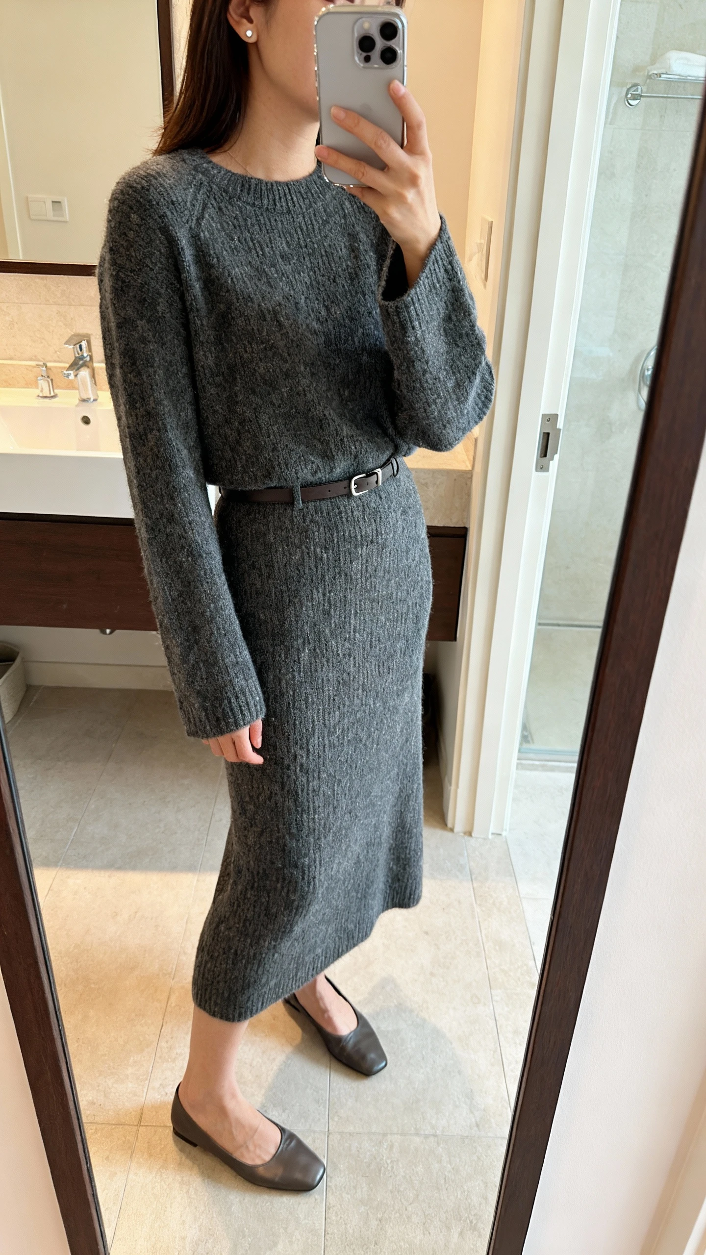 iPhone side-angle mirror selfie of a woman wearing an elevated matching knit set (long-sleeve knit top and midi knit skirt), minimalist belt, sleek flats, and delicate stud earrings, face not visible, bathroom mirror, soft indoor lighting, iPhone photo quality.