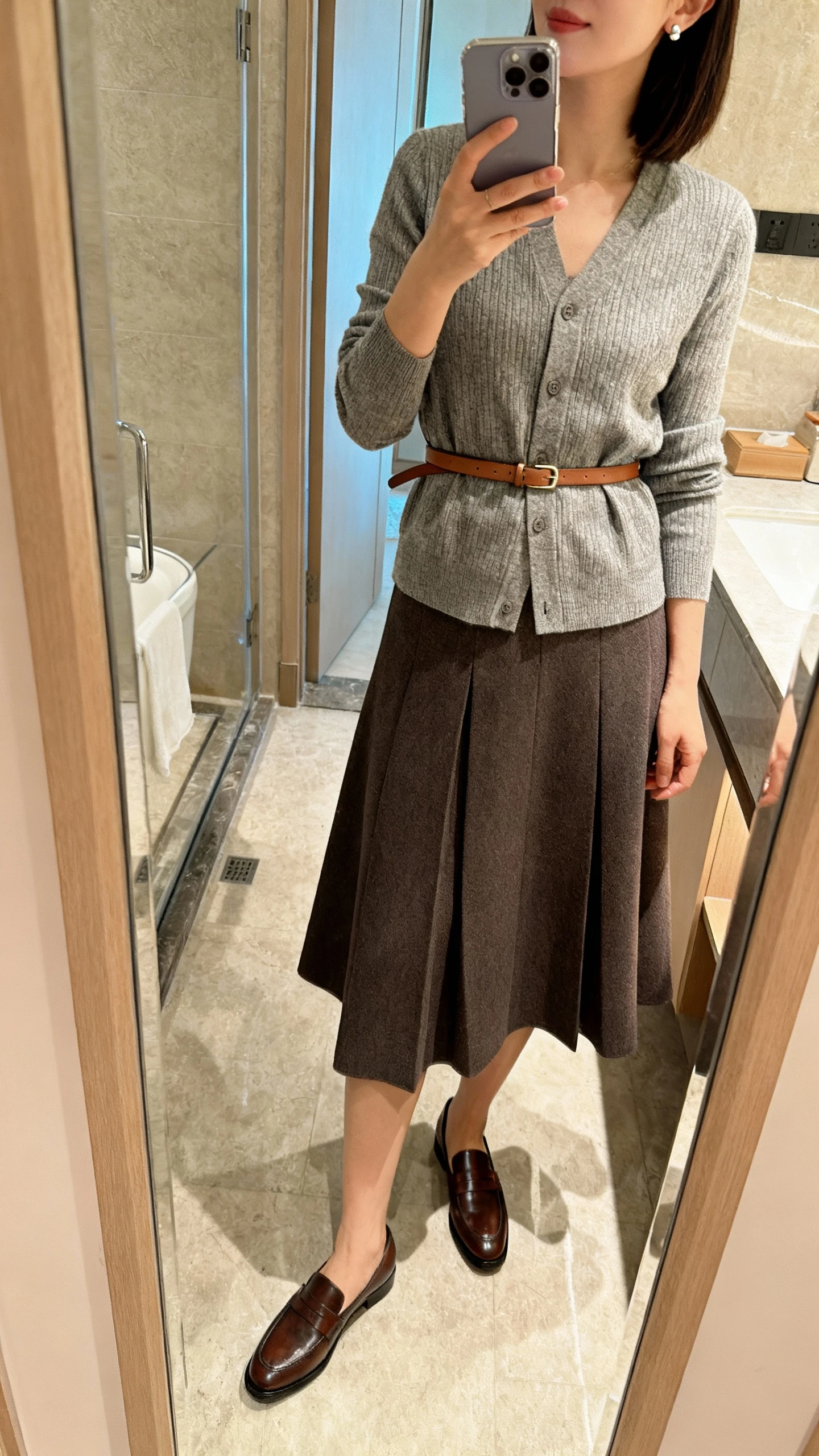 iPhone side-angle mirror selfie of a woman wearing a tailored A-line skirt, belted cardigan cinched at the waist, minimal stud earrings, classic leather loafers, and a slim leather belt, face not visible, bathroom full-length mirror, morning indoor lighting, iPhone photo quality.