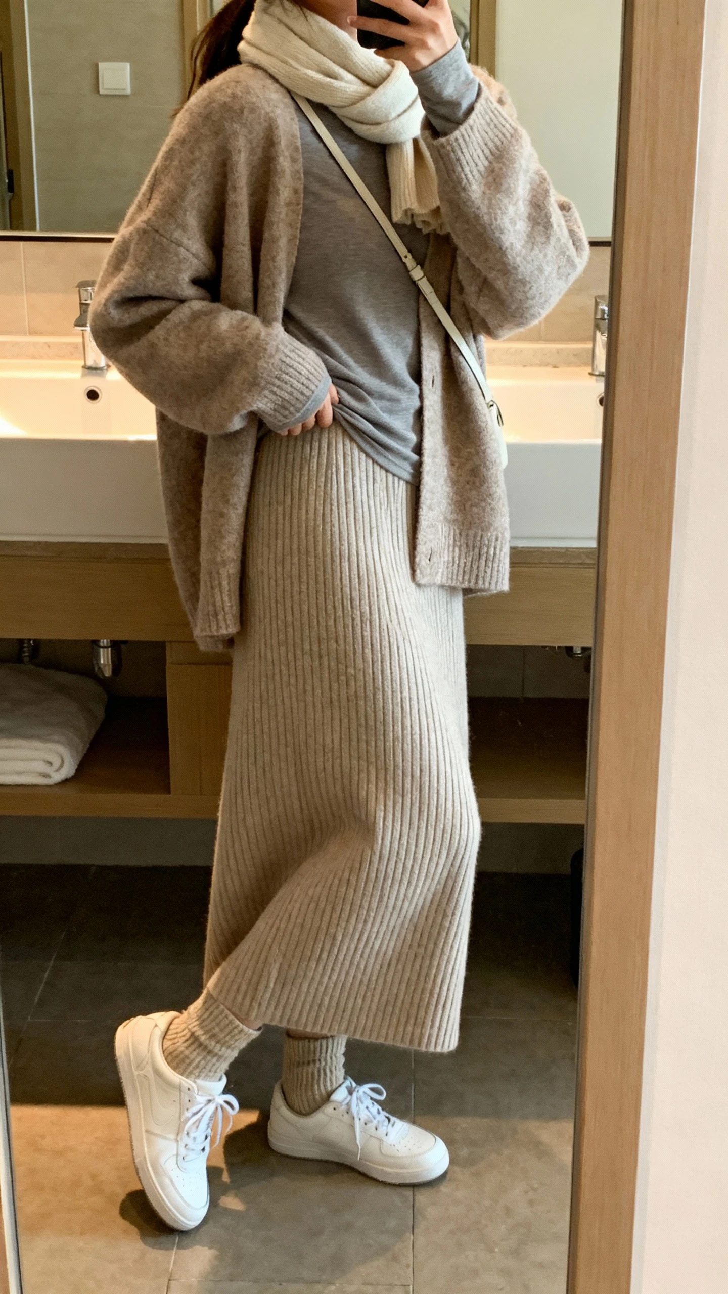 iPhone side-angle mirror selfie of a woman wearing a cozy ribbed knit maxi skirt, soft oversized cardigan layered over a fitted long-sleeve tee, wool socks with clean white sneakers, lightweight scarf, and a minimal crossbody, face not visible, bathroom mirror, warm indoor lighting, iPhone photo quality.