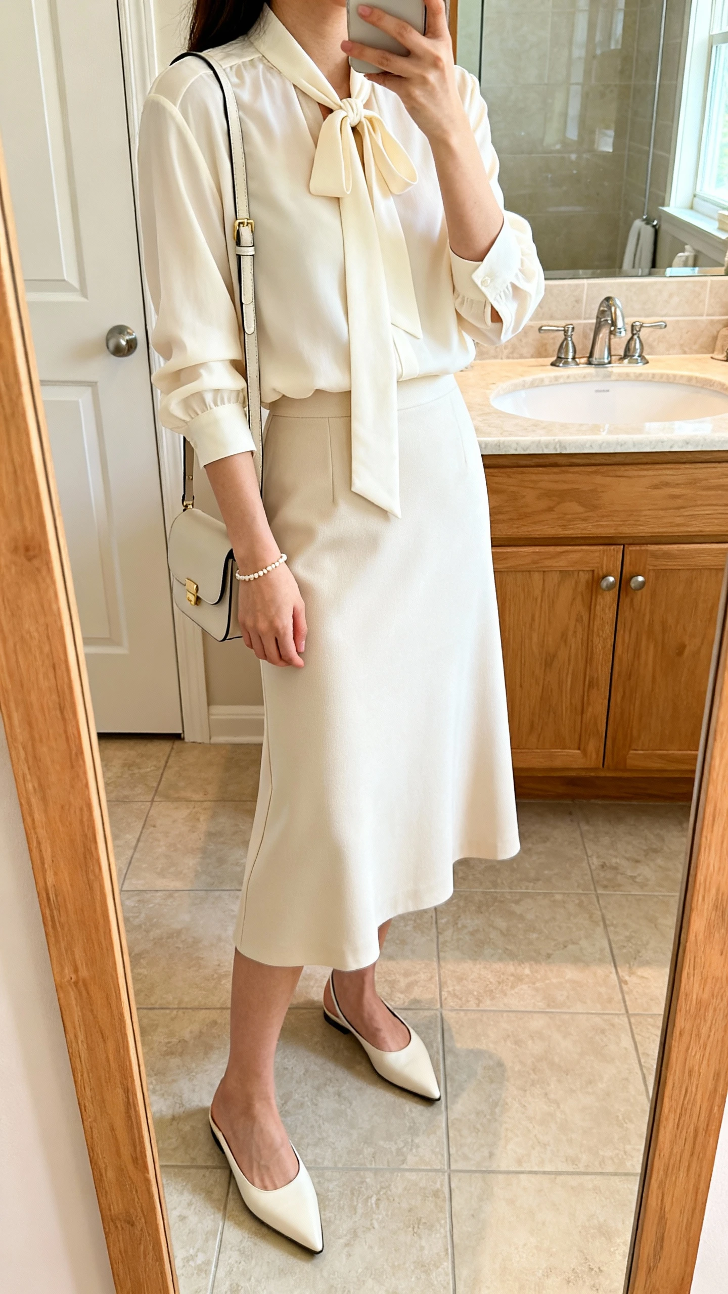 iPhone side-angle mirror selfie of a woman wearing a cream tie-neck blouse paired with a midi A-line skirt, pointed flats, delicate bracelet, and a small shoulder bag, face not visible, cropped at shoulders, bathroom mirror, soft morning light, iPhone photo quality.