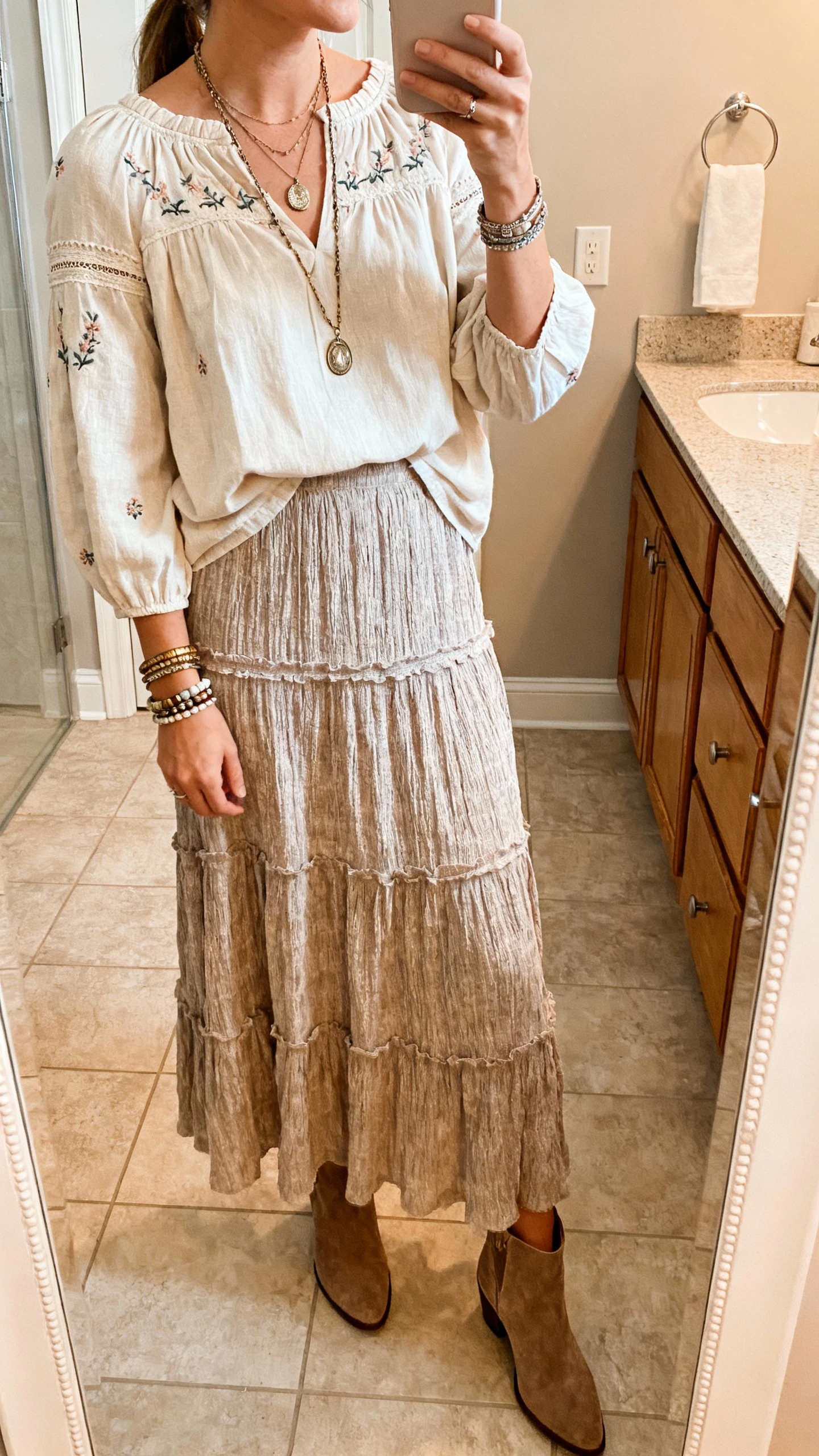 iPhone side-angle mirror selfie of a woman wearing a peasant blouse with subtle embroidery paired with a tiered textured maxi skirt, layered pendant necklaces, stackable bracelets, and suede ankle boots, face not visible, bathroom mirror, warm indoor lighting, iPhone photo quality.