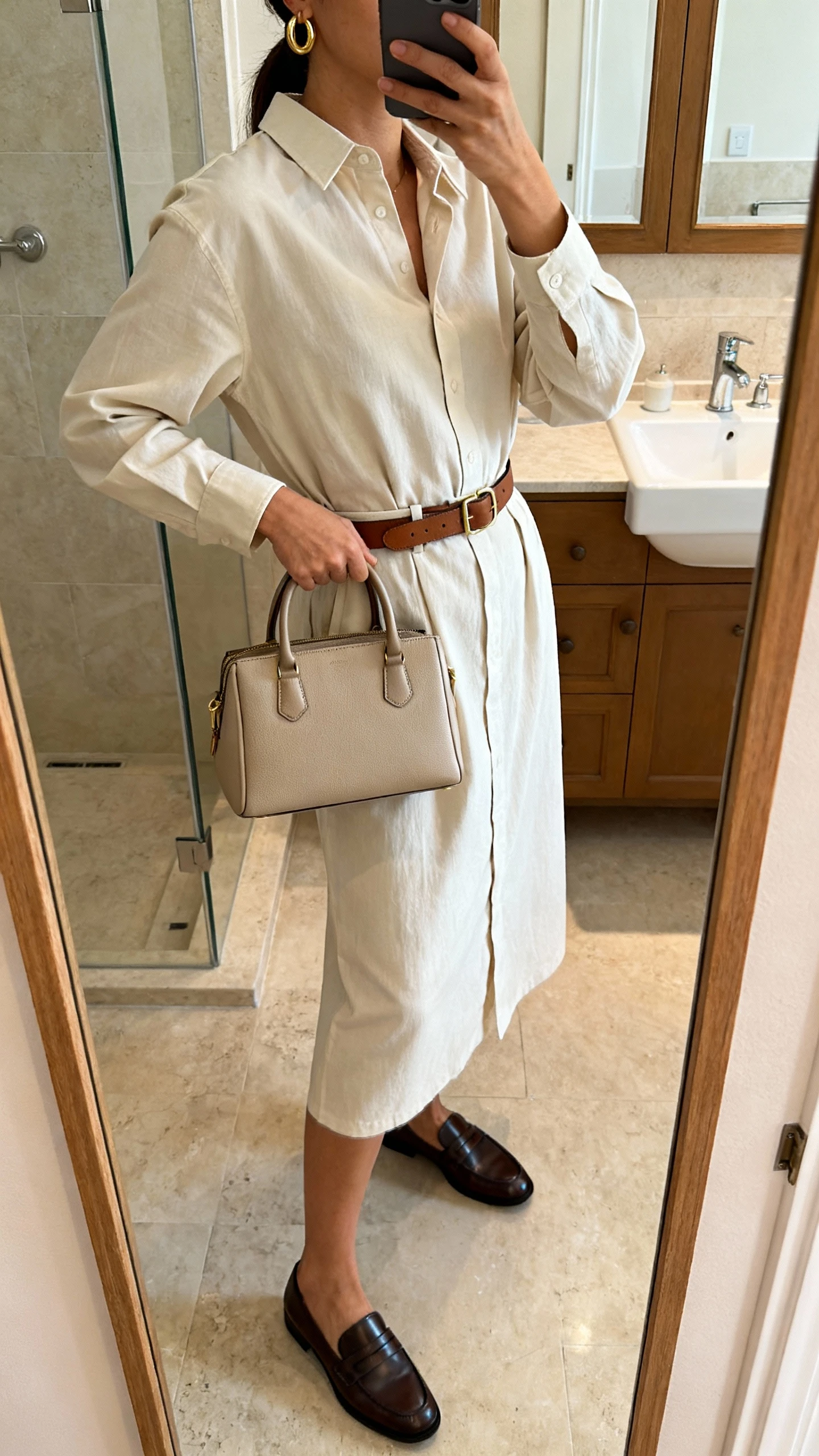 iPhone side-angle mirror selfie of a woman wearing a belted shirt dress with quiet luxury vibes, minimal gold hoop earrings, slim leather belt, structured top-handle bag, and sleek loafers, face not visible, bathroom full-length mirror, morning indoor lighting, iPhone photo quality.