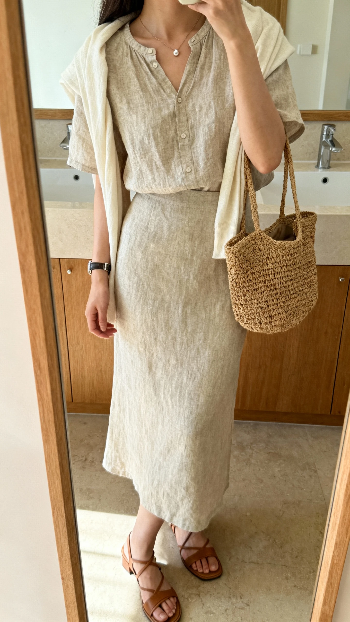 iPhone side-angle mirror selfie of a woman wearing a short-sleeve linen blouse with a modest neckline and a matching linen midi skirt, strappy low-heel sandals, a woven straw bag, a minimal pendant necklace, small studs, a watch, and a lightweight shawl over her shoulders; face not visible, bathroom mirror, soft morning light, iPhone photo quality.